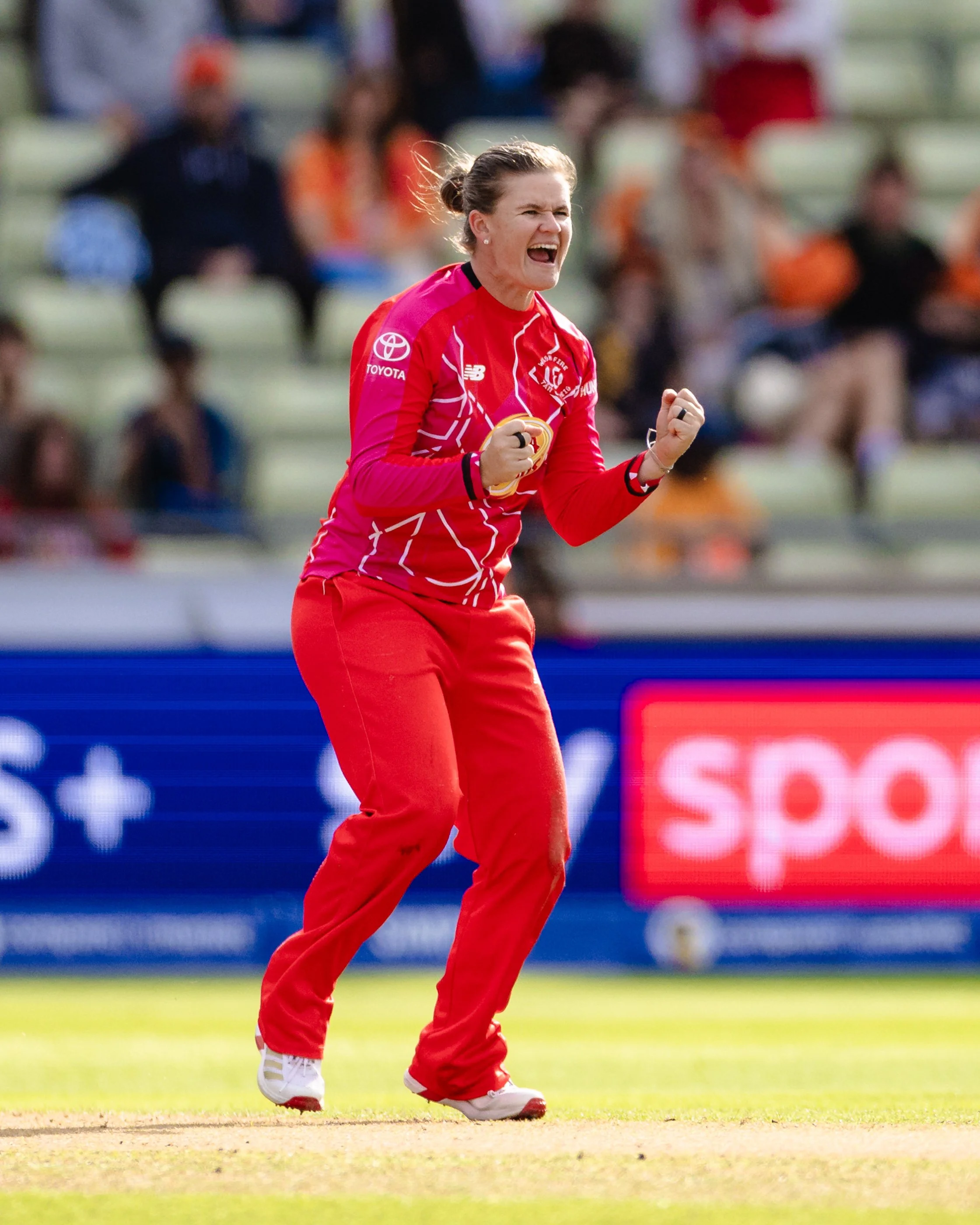 Celebrating a victory during a cricket match, a woman in a red sports uniform with white and pink accents clenches her fists and shouts joyfully on the field, with a blurred crowd in the background.