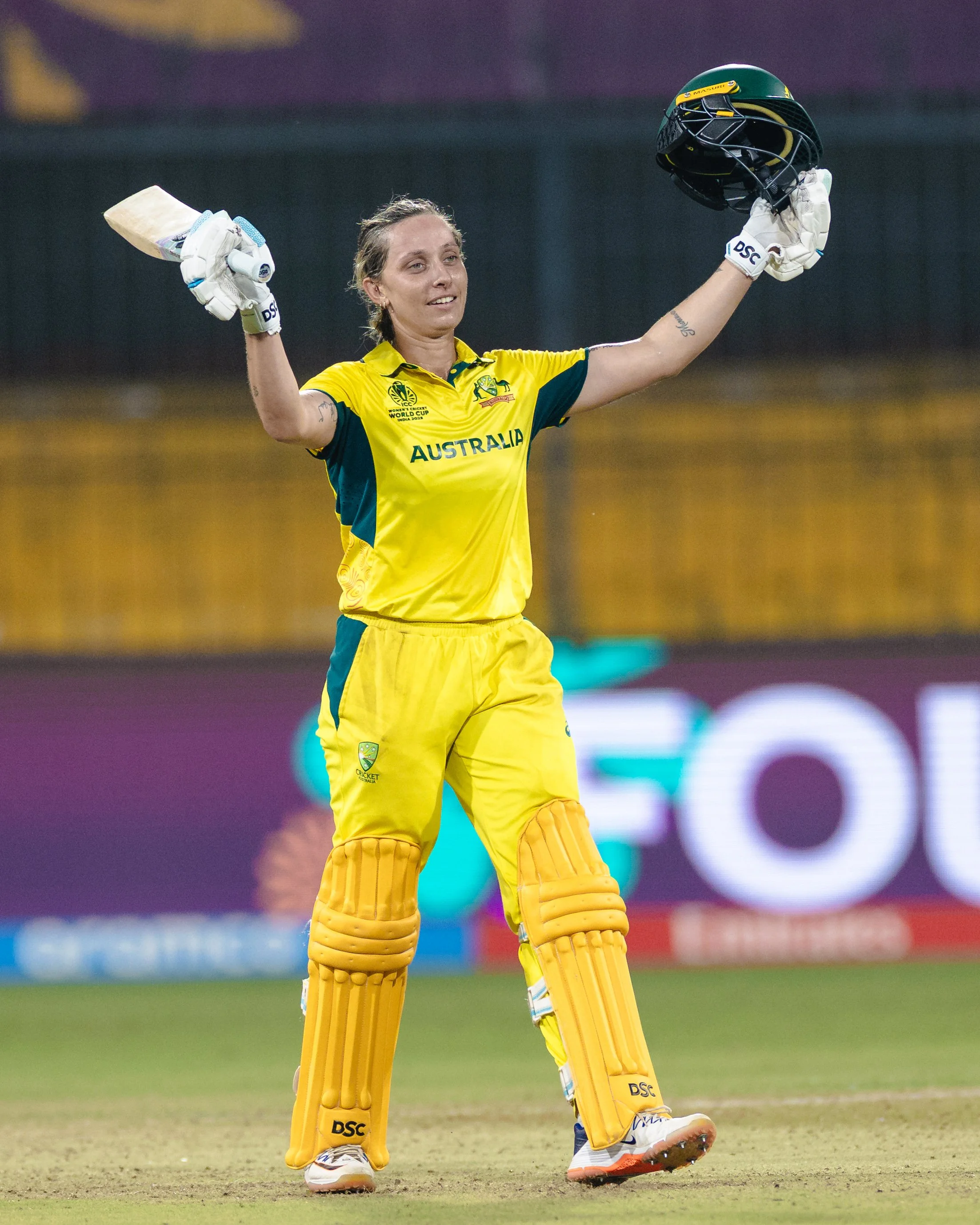 A female cricket player in yellow uniform holding a cricket helmet and a bat, smiling, on a cricket field.
