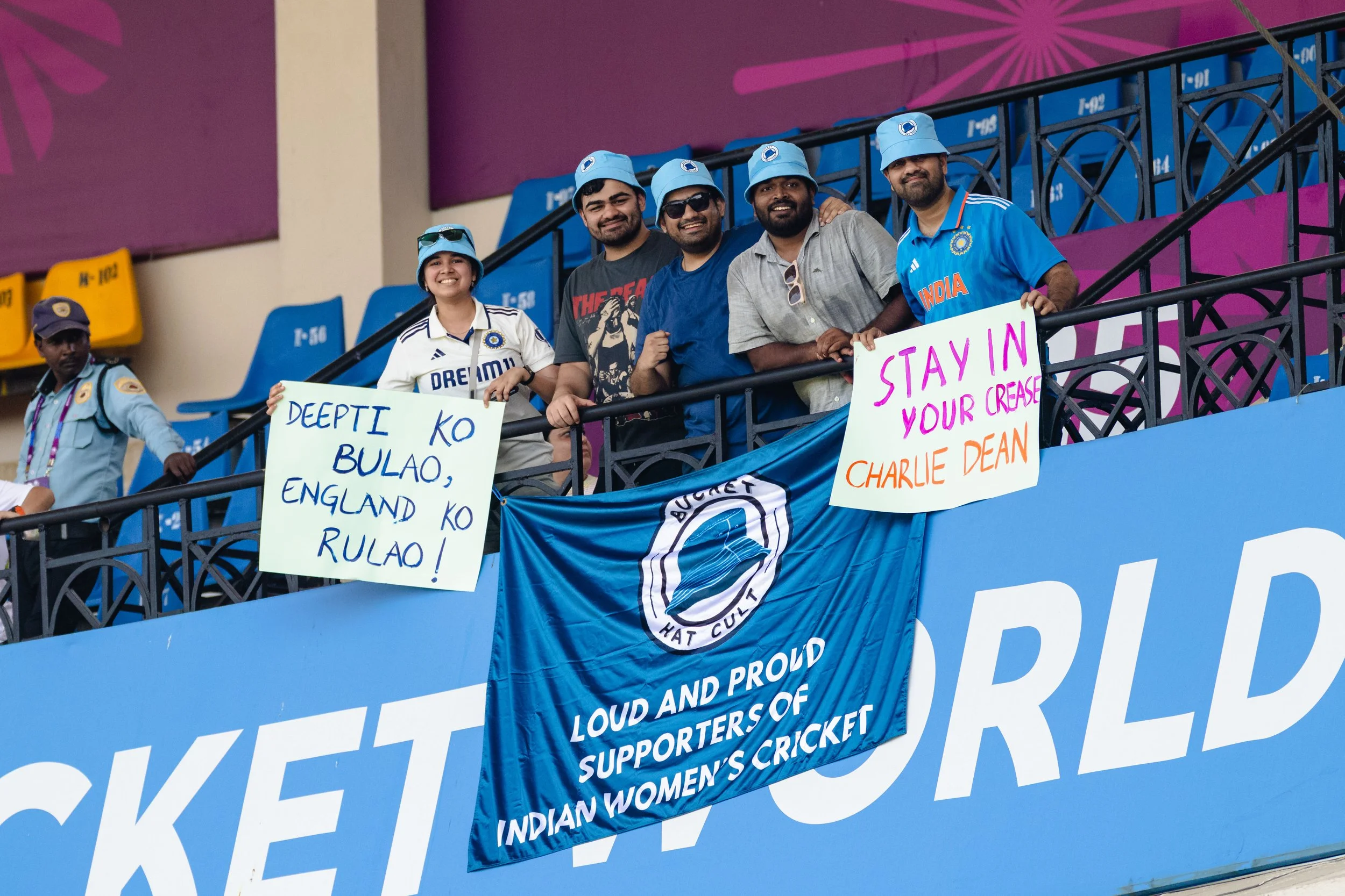 A group of cricket supporters at a stadium, holding signs and wearing blue hats, with one person in an Indian cricket jersey. The stadium seats are visible in the background.