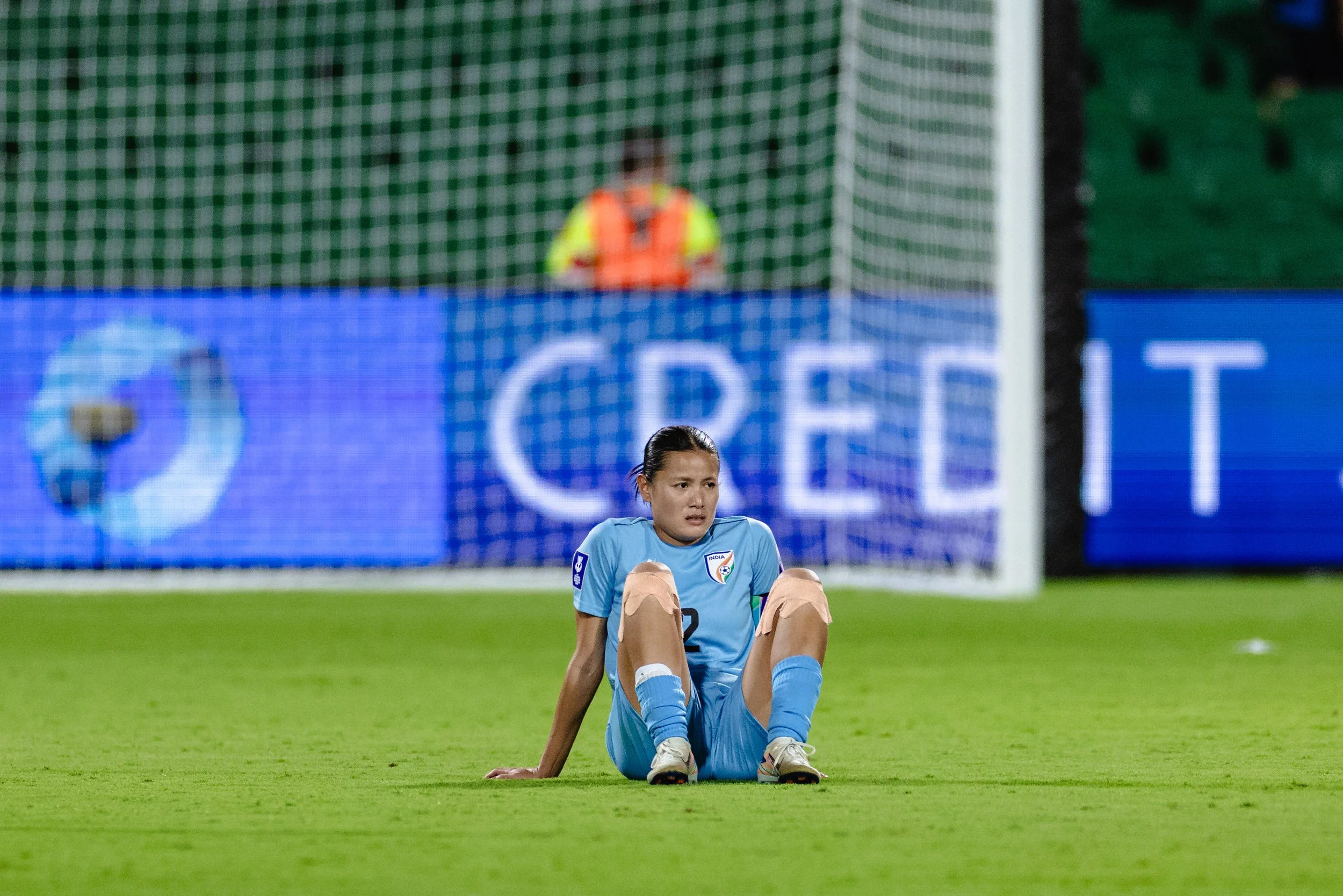 A female soccer player sitting on the field, wearing a blue uniform. She appears distressed or tired, with a soccer goal and a security guard in the background.