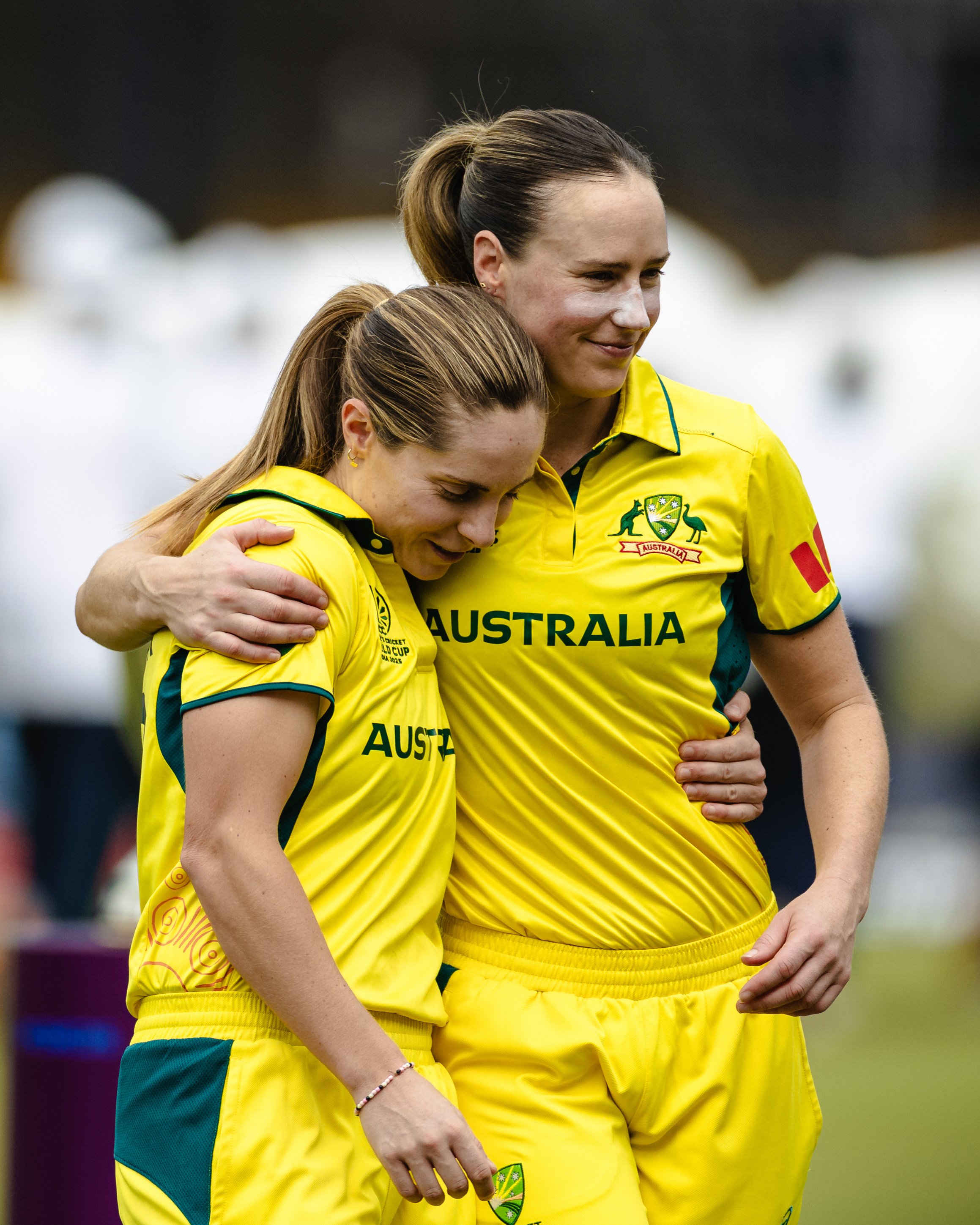 Two Australian female cricket players wearing yellow jerseys hugging each other on the field, one has her head bowed while the other gazes forward with a smile.