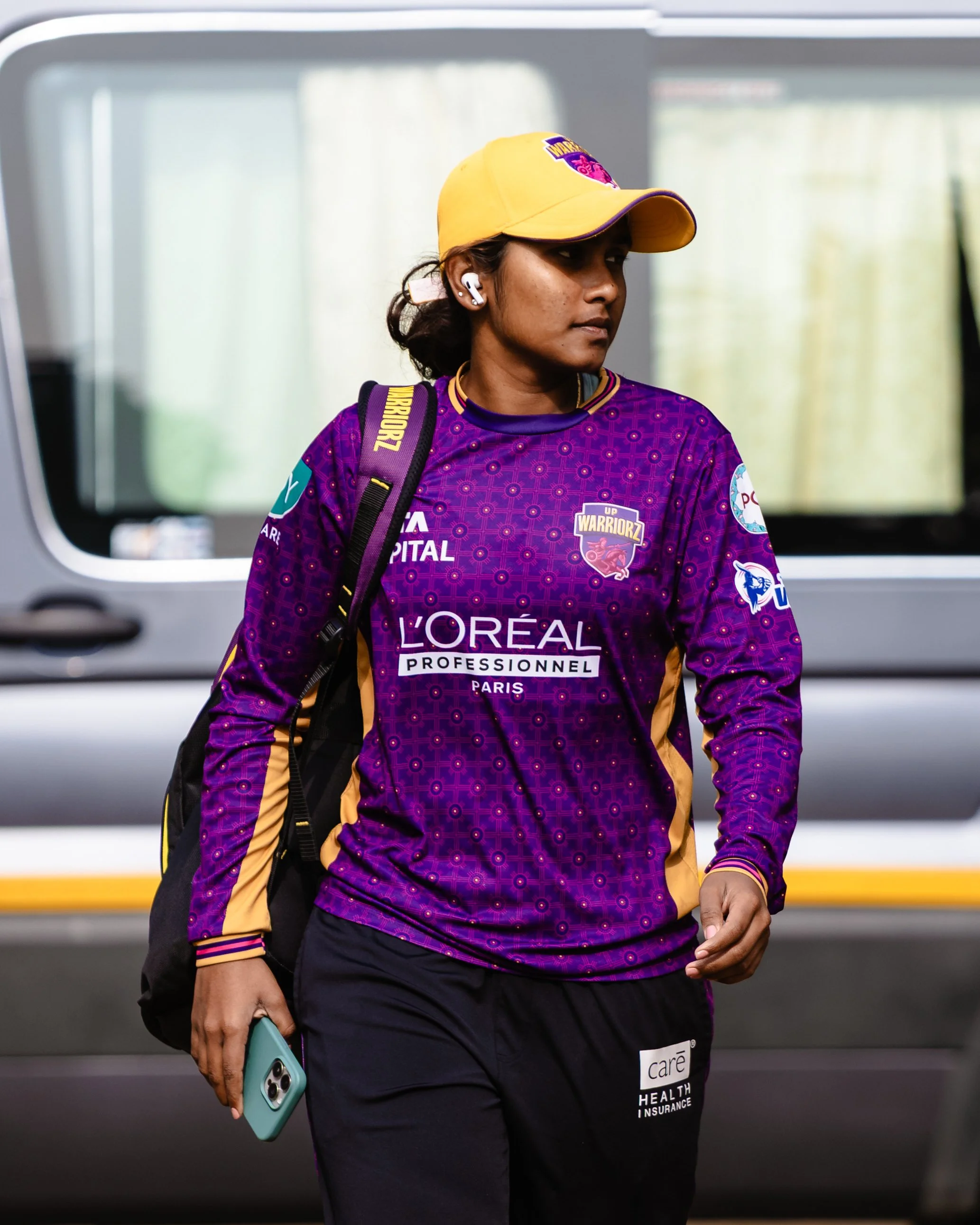 A woman wearing a yellow cap and purple sports jersey with logos, carrying a black backpack, and holding a teal phone, walking near a window in a transportation station.