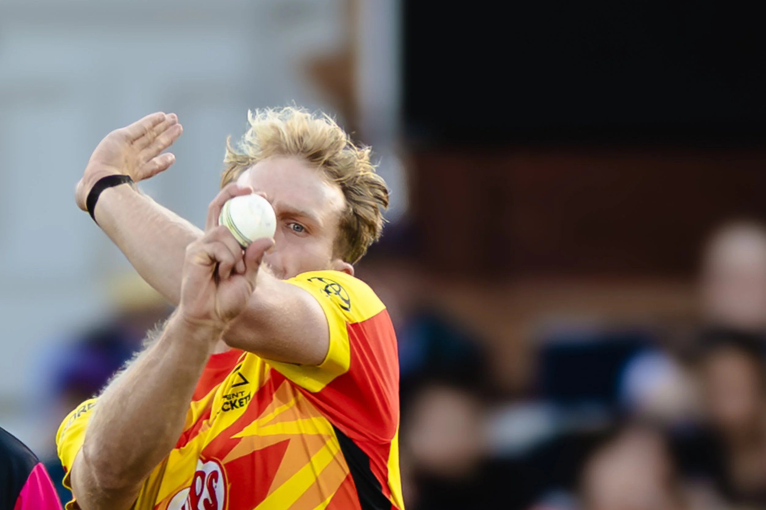 A cricket player in a yellow and red jersey is preparing to bowl during a game, with their right arm raised and holding a cricket ball.