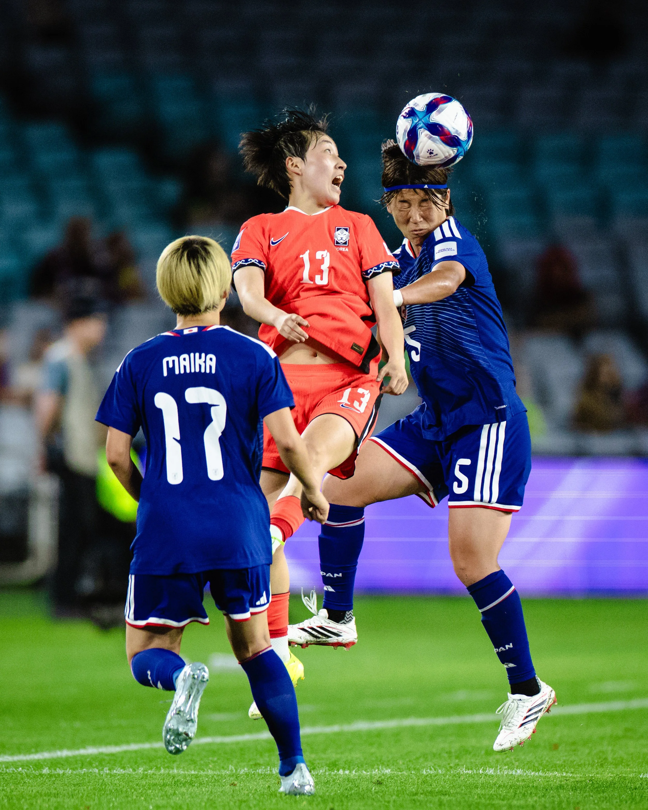Two female soccer players from different teams jump to head the ball during a match, with a third player observing. The player in red is wearing jersey number 13, and the player in blue is wearing jersey number 5.
