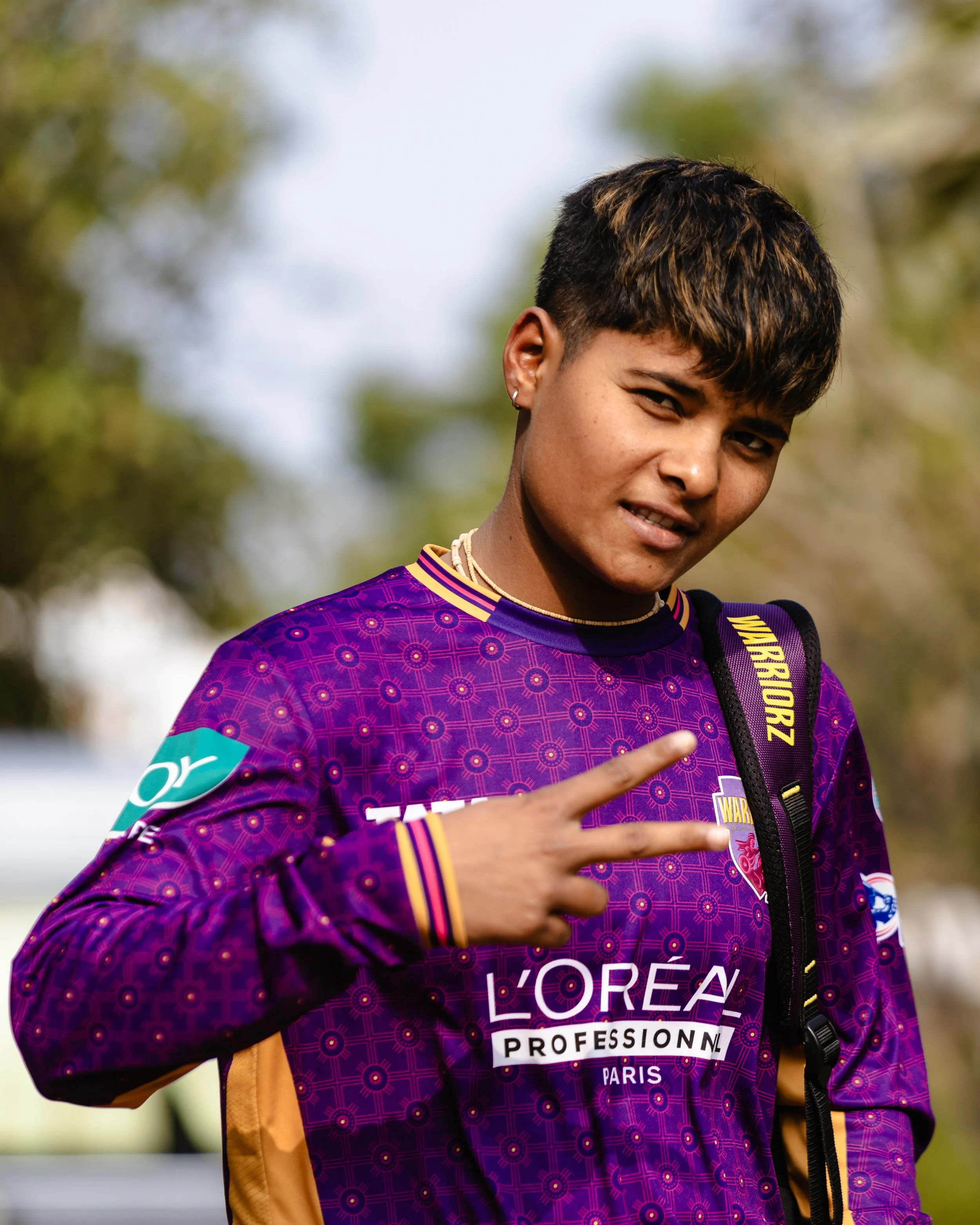 Close-up of a young person with short dark hair, sporting a purple sports jersey with gold and black accents, and a necklace, outdoors with blurred trees in the background, making a hand gesture.
