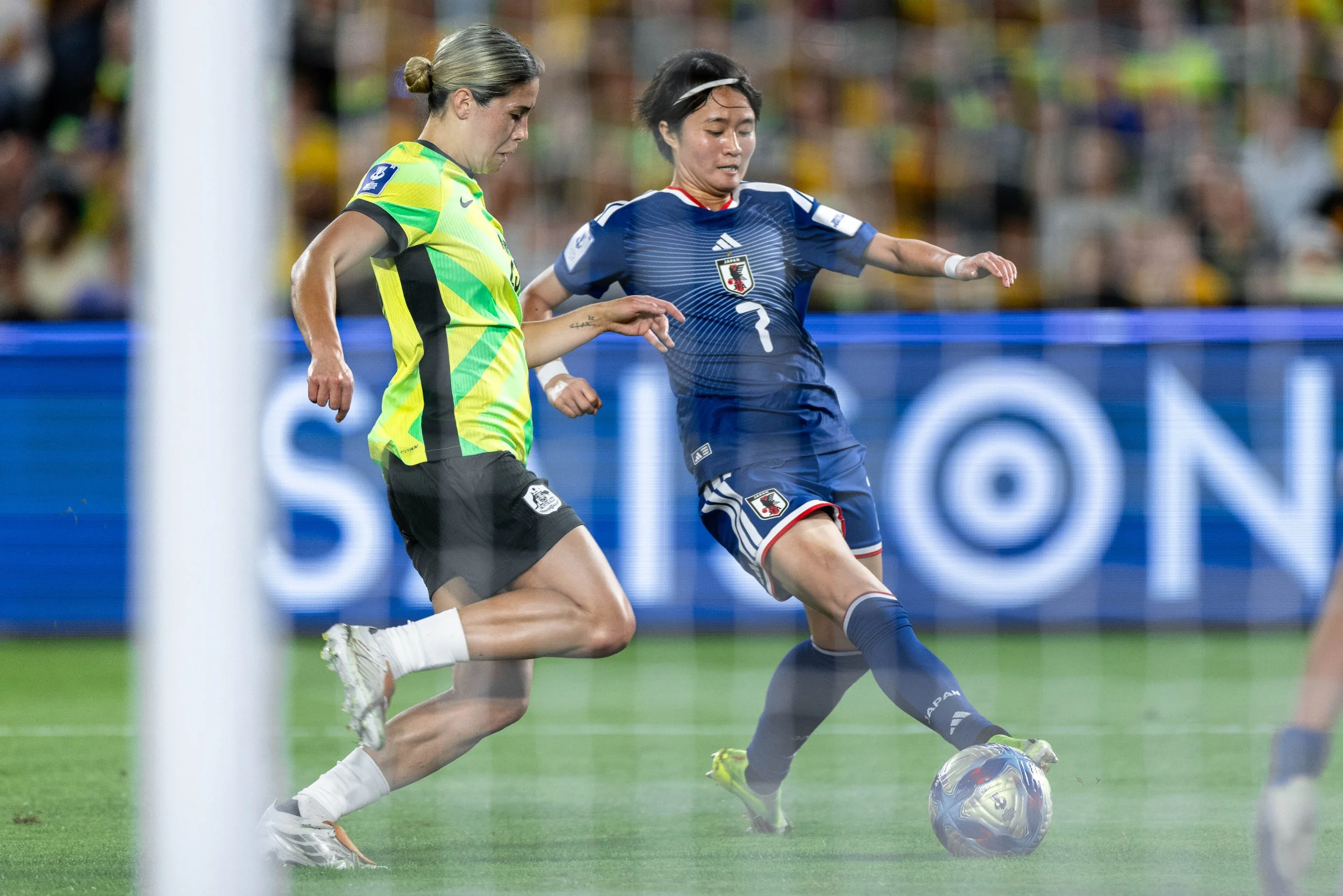 Two female soccer players competing for the ball on the field during a match. One player wears a yellow and green jersey, and the other wears a blue jersey with a Japan emblem. The scene is captured mid-action with a crowd in the background.