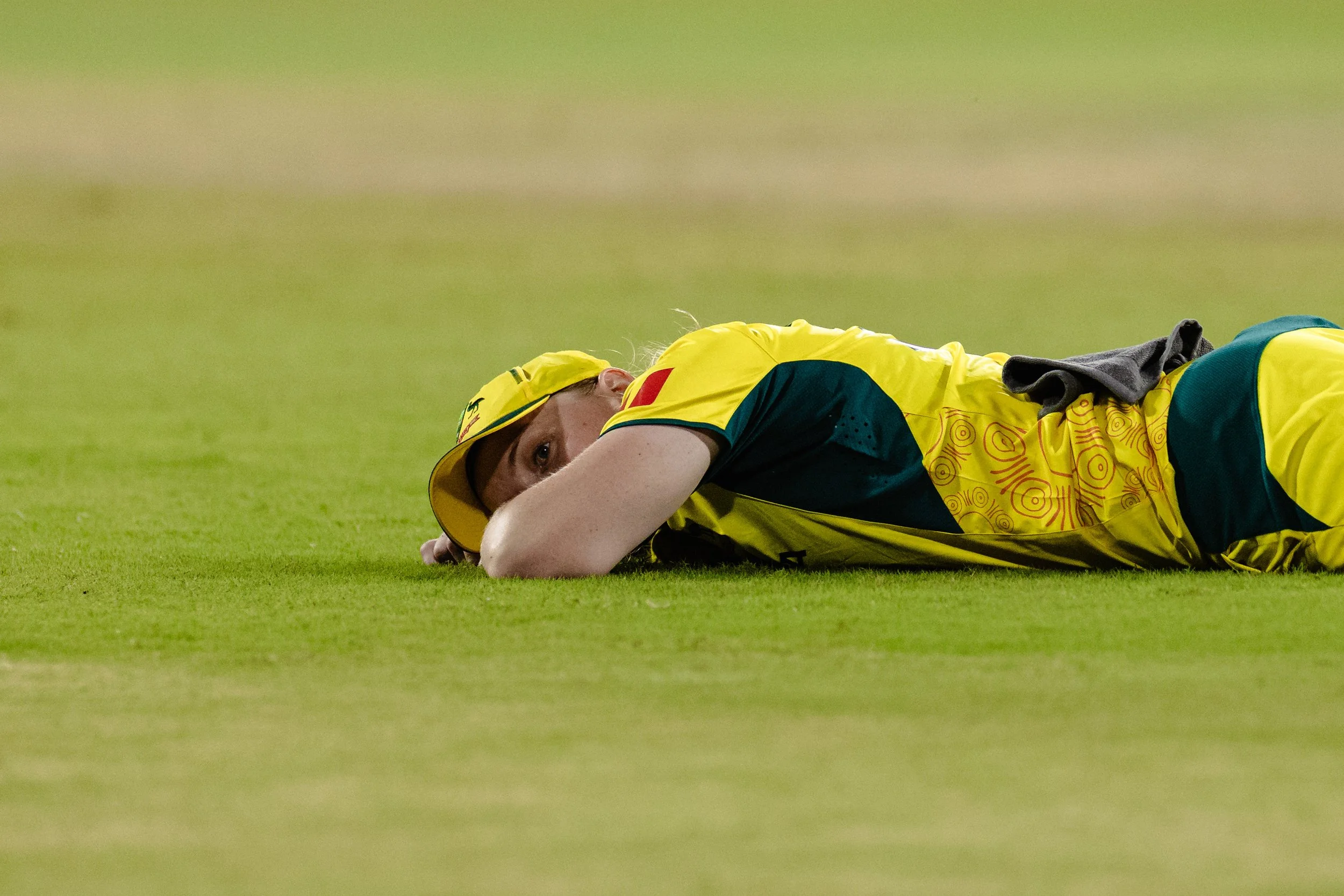 An athlete in a yellow uniform lying on the grass field, looking at the camera, with a gray towel on their back.