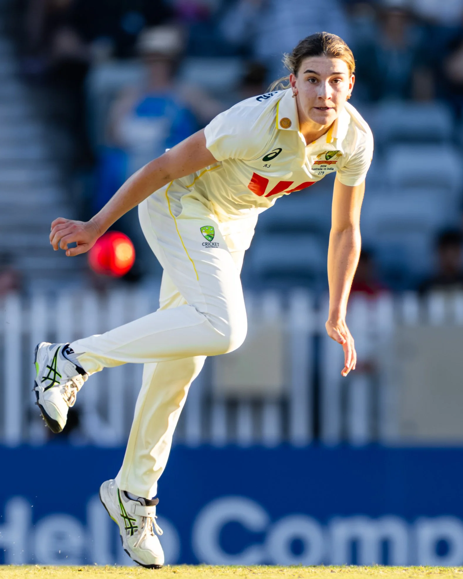 A female cricket player in a white uniform appears to be bowling during a match, with blurred spectators in the background.
