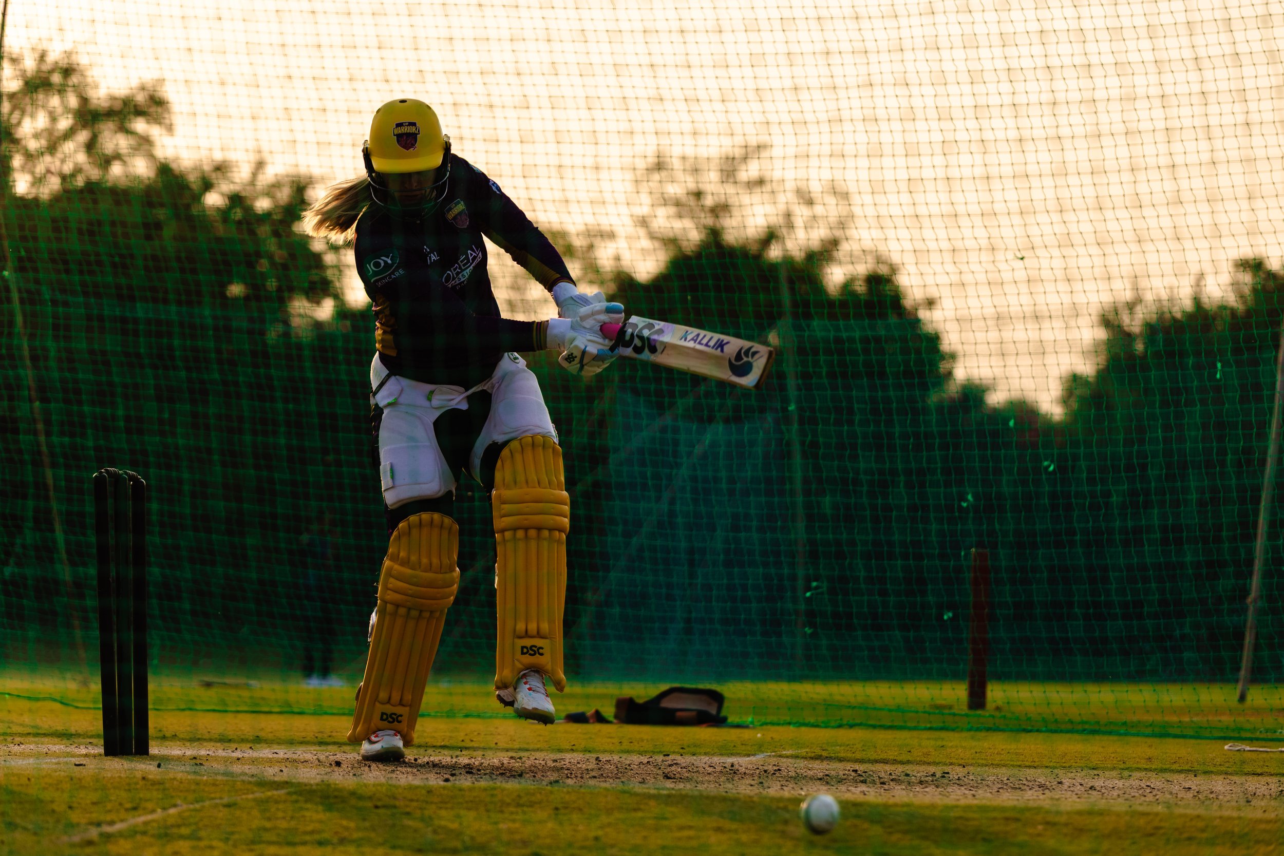 Cricketer practicing during sunset, wearing a helmet, pads, and uniform, with a cricket ball on the ground.