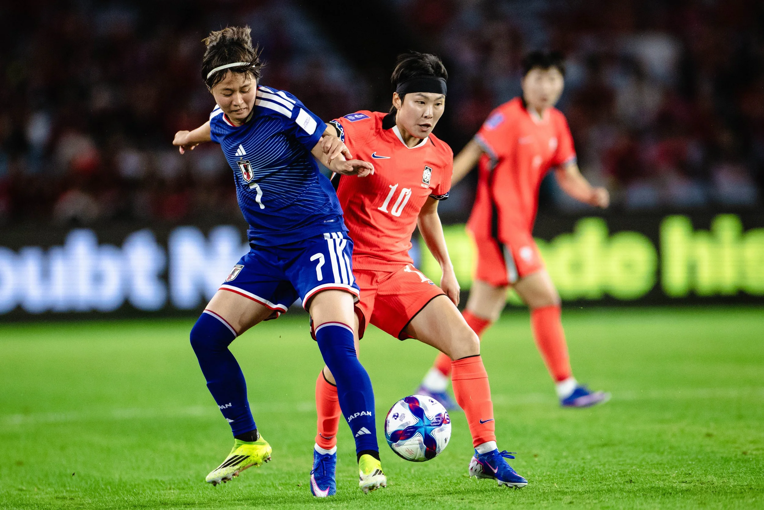 Two female soccer players, one in a blue jersey and the other in a red jersey, contest for the ball during a match on a green field at night.