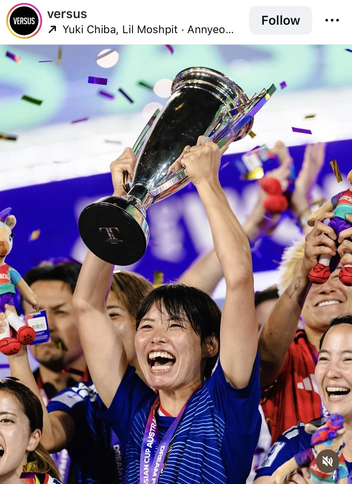 A woman holding a large silver trophy celebrating victory with a big smile, surrounded by teammates and confetti, at a sports event.
