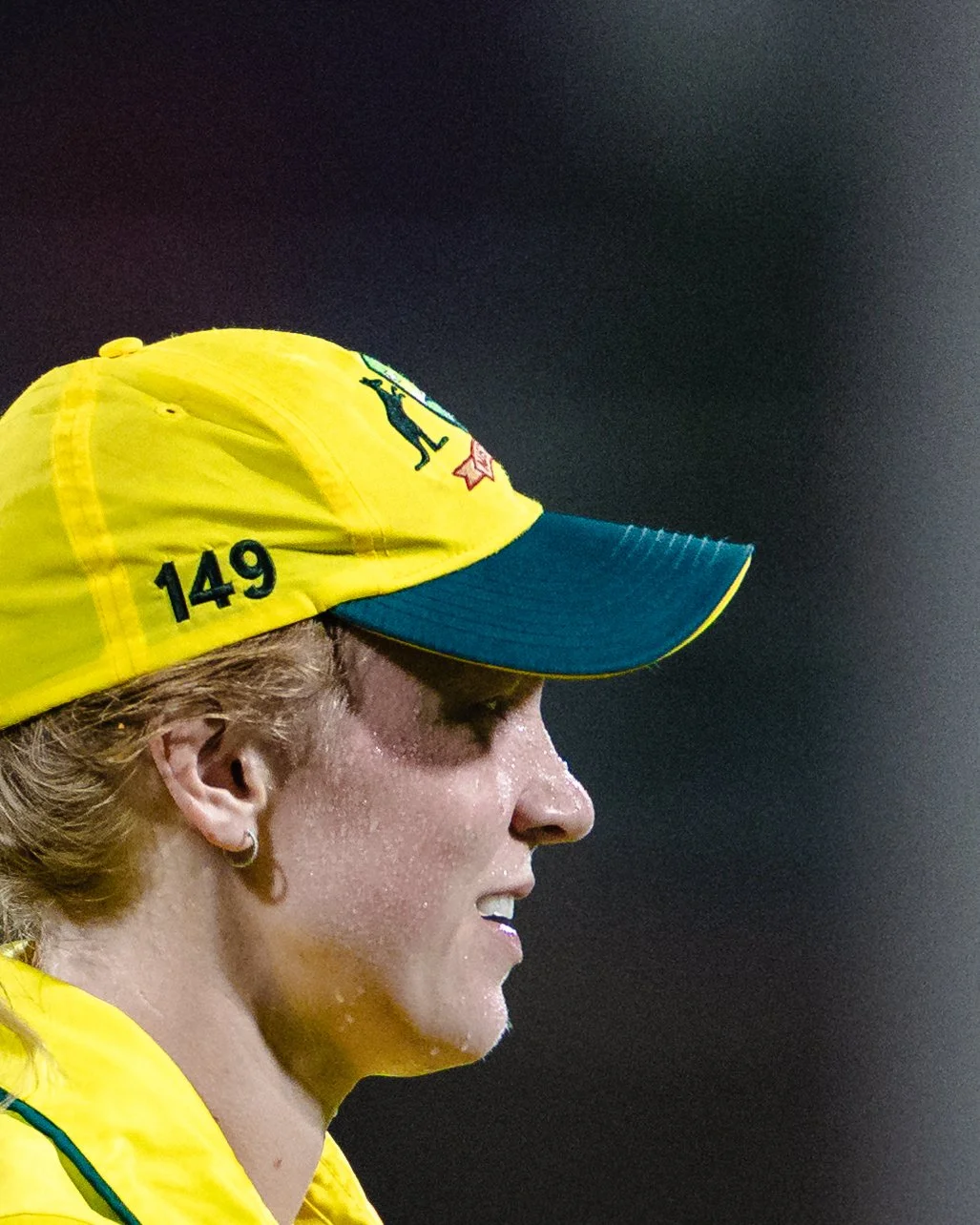 A side profile of a woman in a yellow and green sports uniform with a yellow cap, wet with sweat.