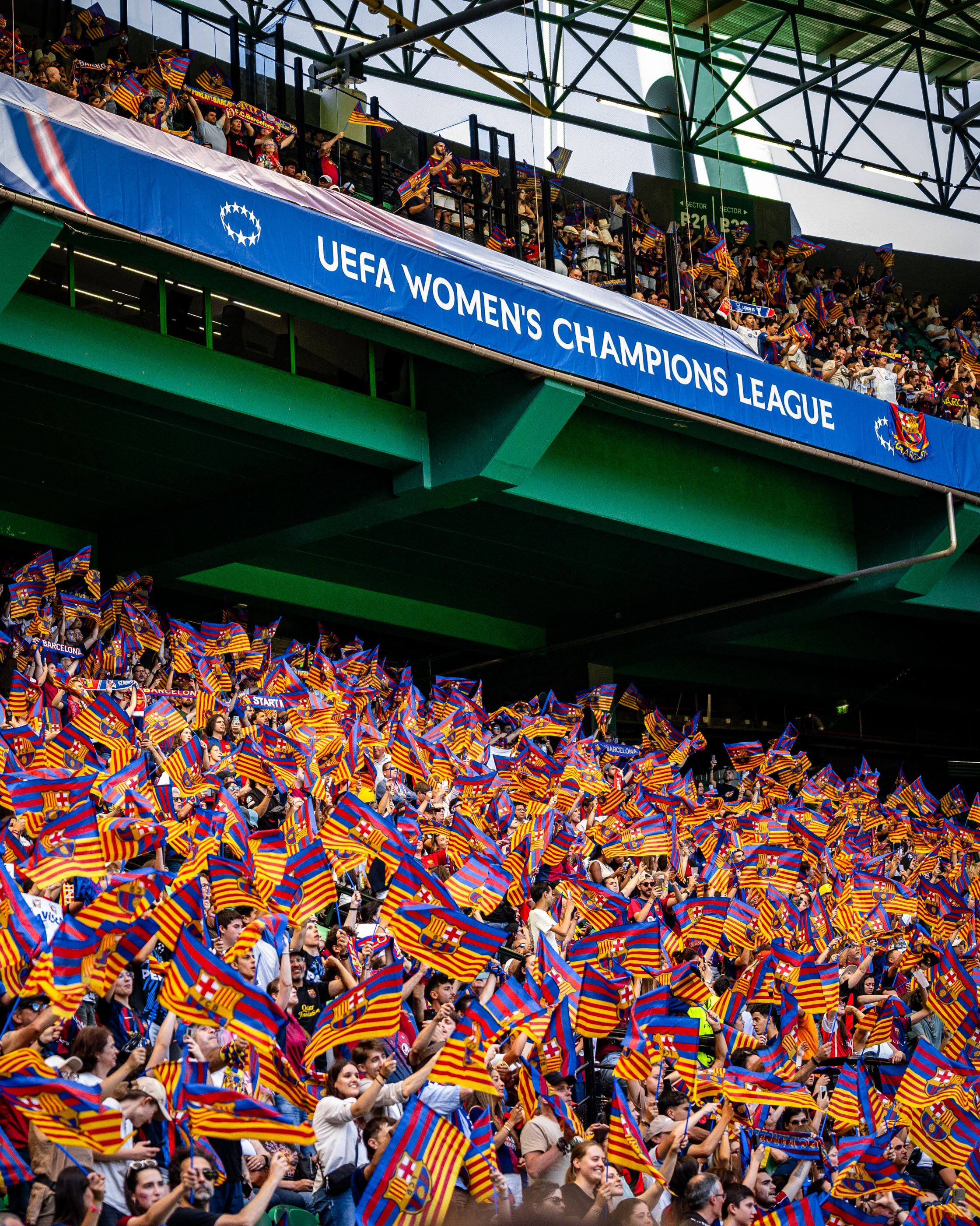 Crowd of football fans in stadium stands waving yellow and red striped flags with a blue cross, celebrating during UEFA Women's Champions League match.