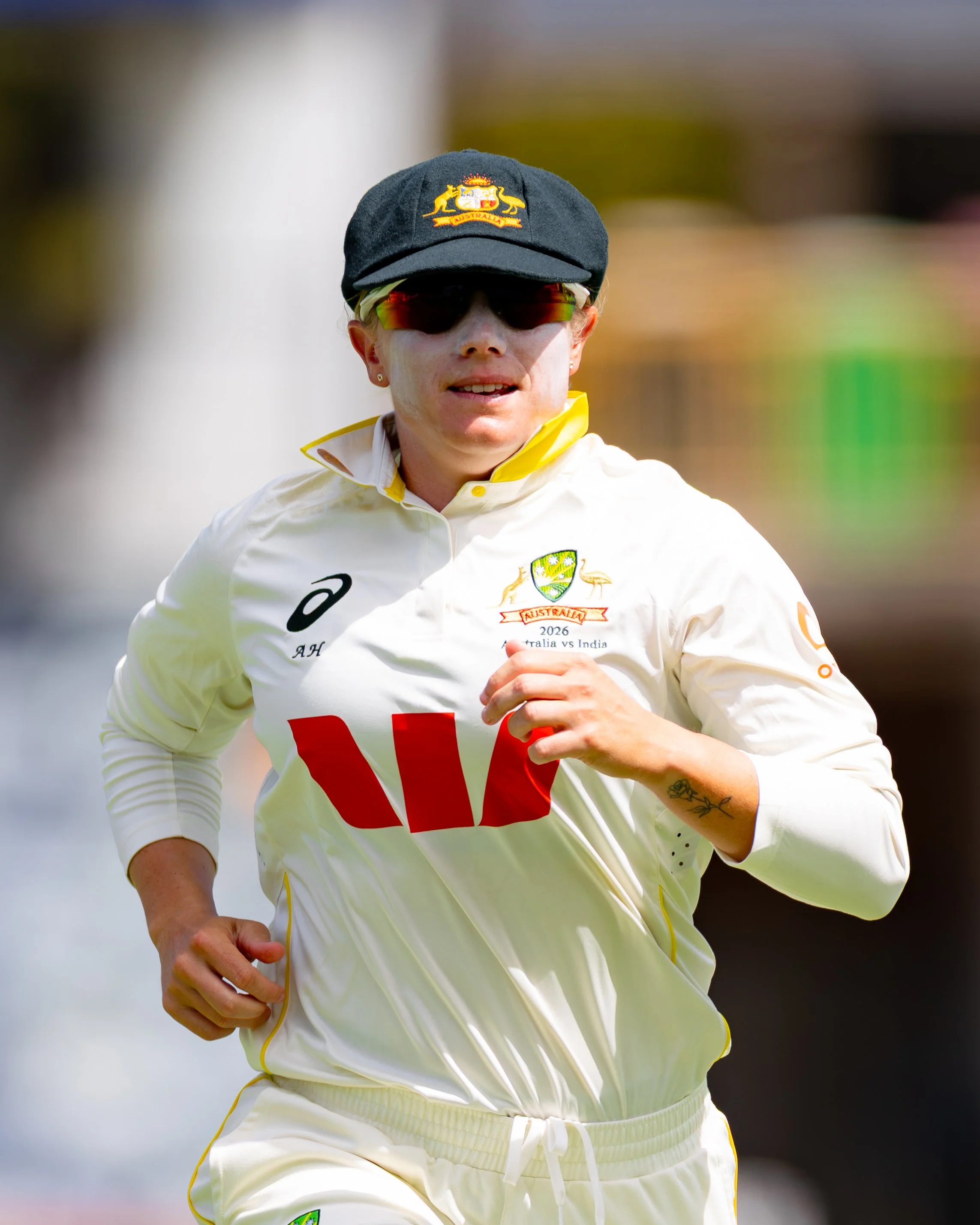 A female cricket player wearing sunglasses and a hat with the Australian emblem is running during a match, with a blurred background.
