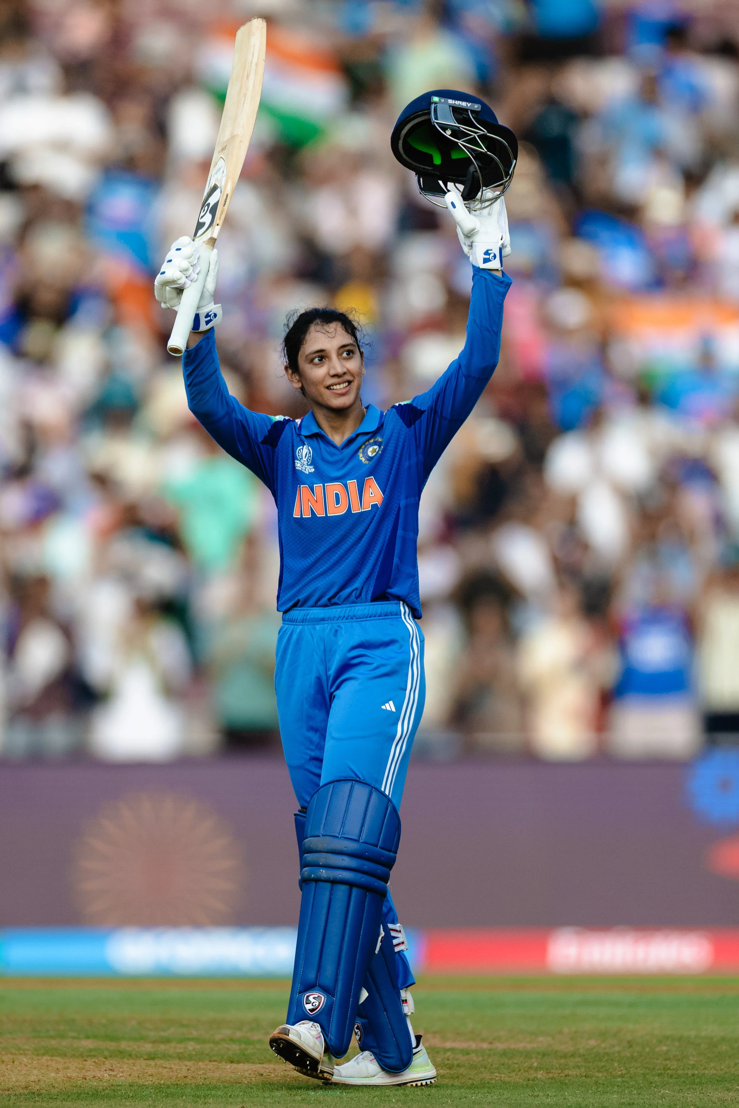 An Indian female cricket player in blue uniform celebrating on a cricket field, holding a helmet in her right hand and a cricket bat in her left hand, with a crowd in the background.