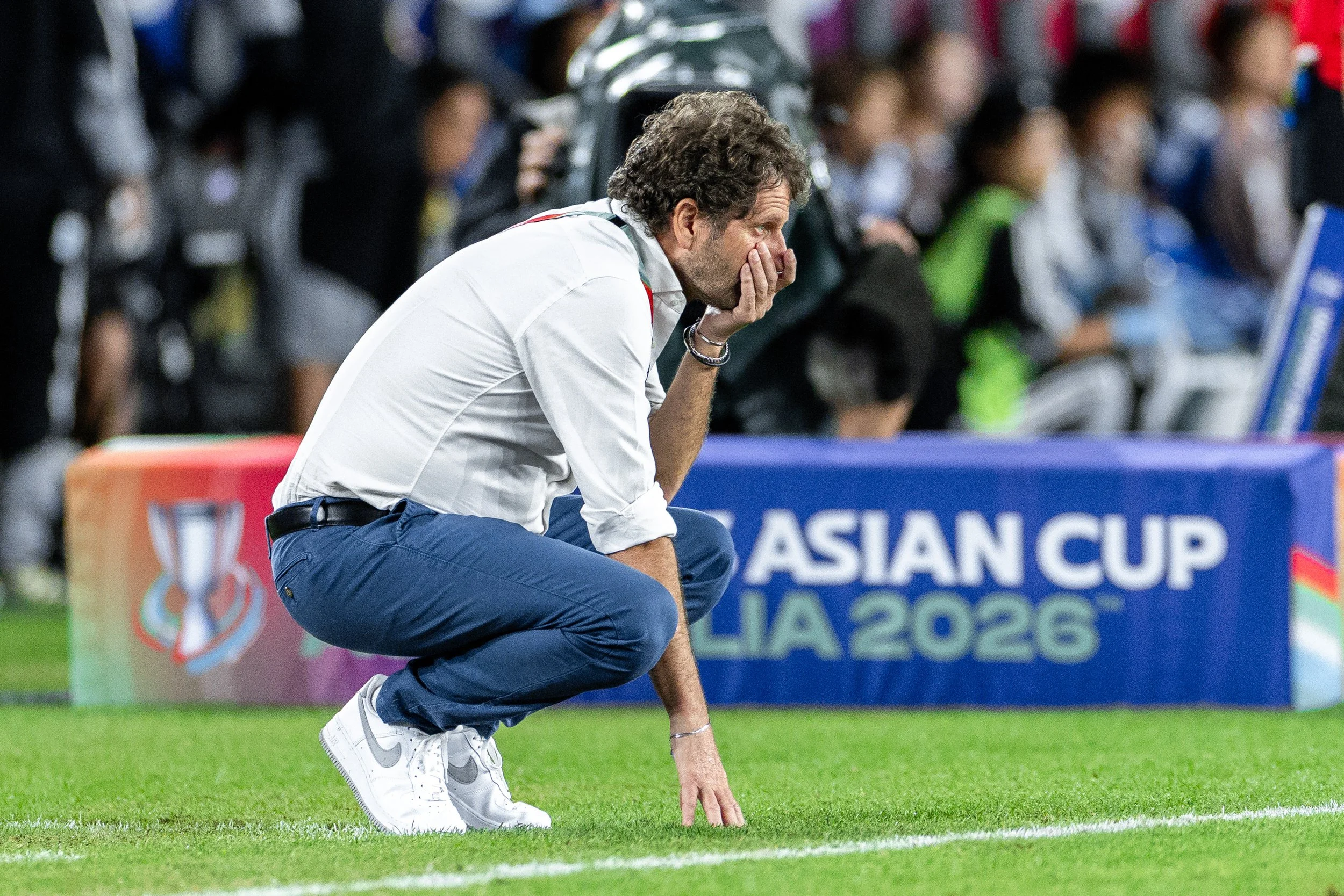 A man with curly hair and a beard, dressed in a white shirt and blue pants, kneeling on the grass with one hand on the ground and the other covering his face, at a sports event with a crowd in the background.