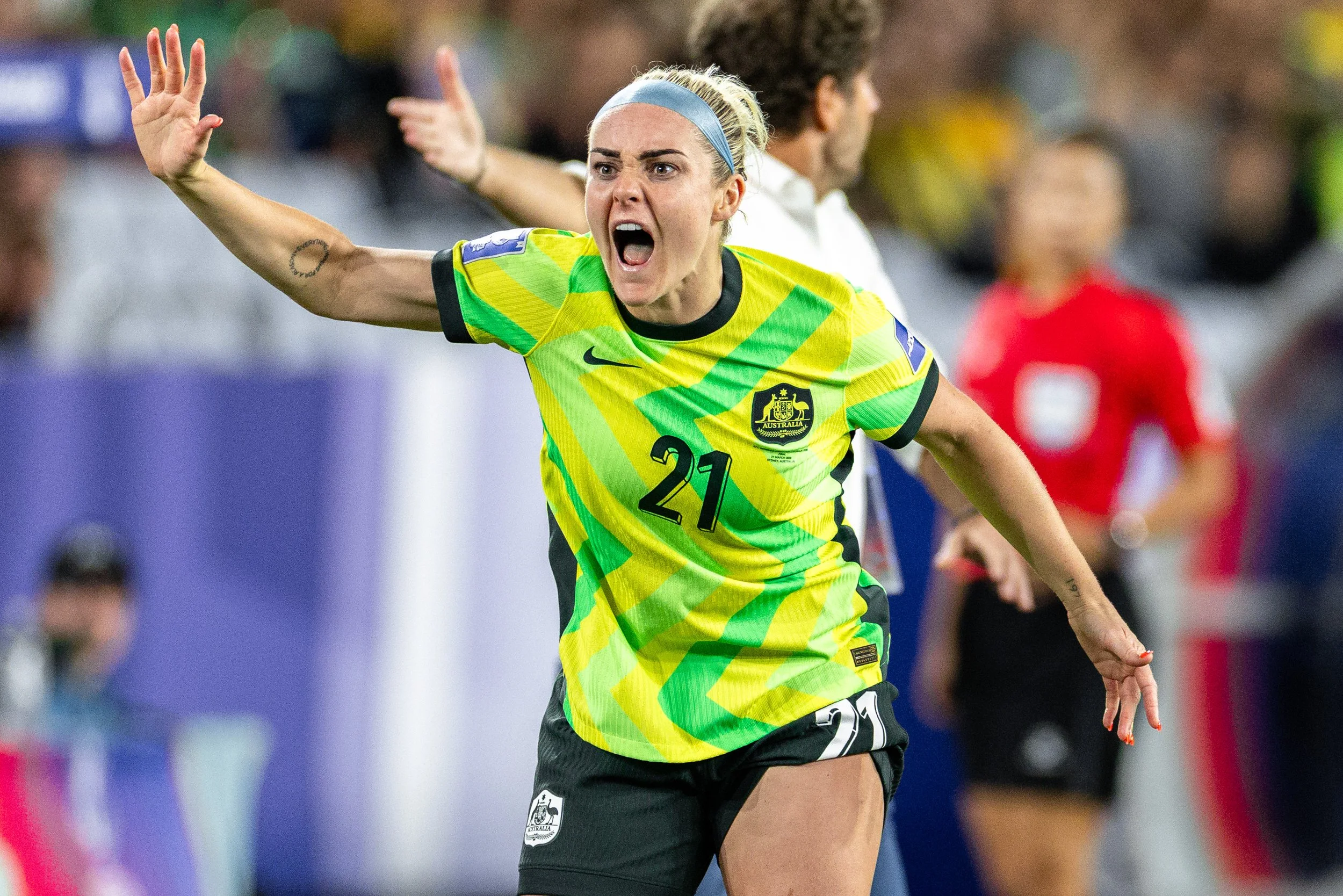 A female soccer player wearing a yellow and green jersey with the Australian emblem, number 21, has her mouth open in a shout or celebration, with her right arm extended and a tattoo on her inner arm. She is on a soccer field with a blurred crowd and