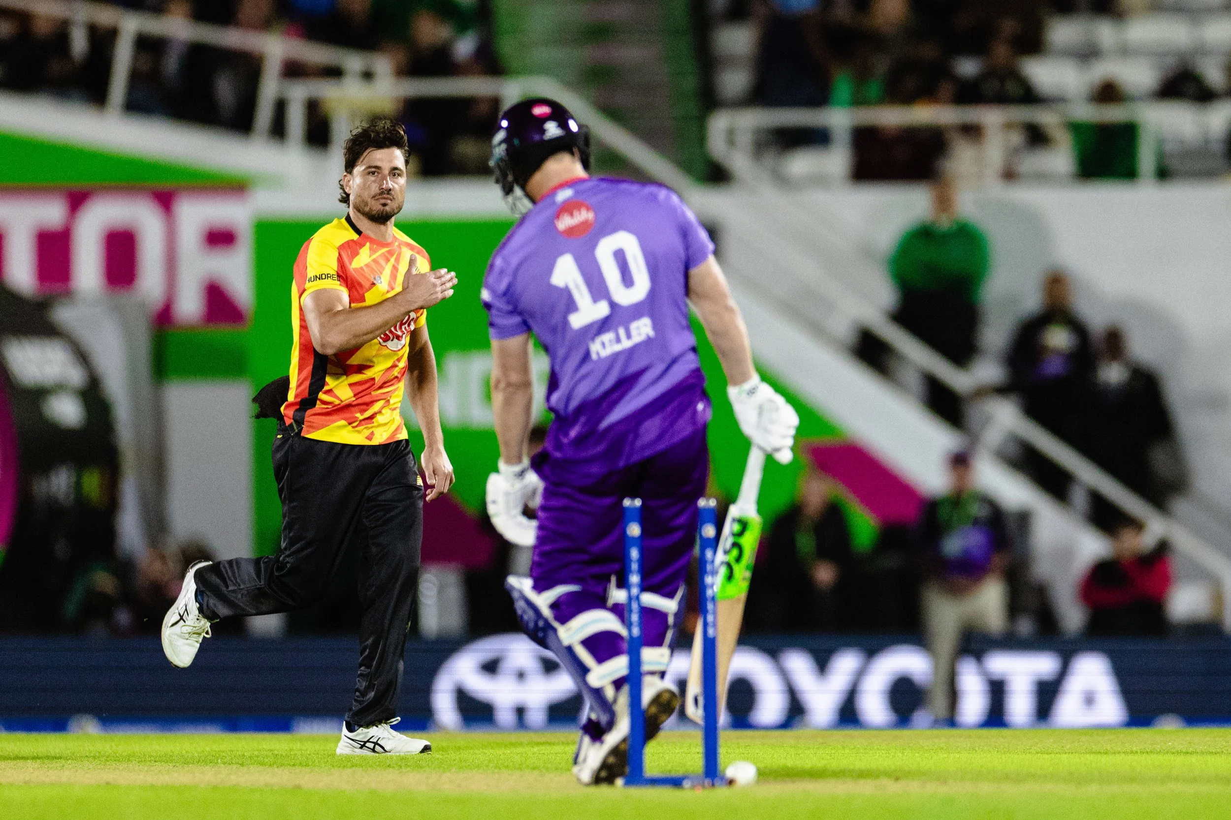 A man in a yellow and red sports jersey appears to be running on a field during a hockey game. Another person, dressed in purple sports attire and wearing a helmet, is facing away from the camera. The scene is set in an indoor stadium with spectators