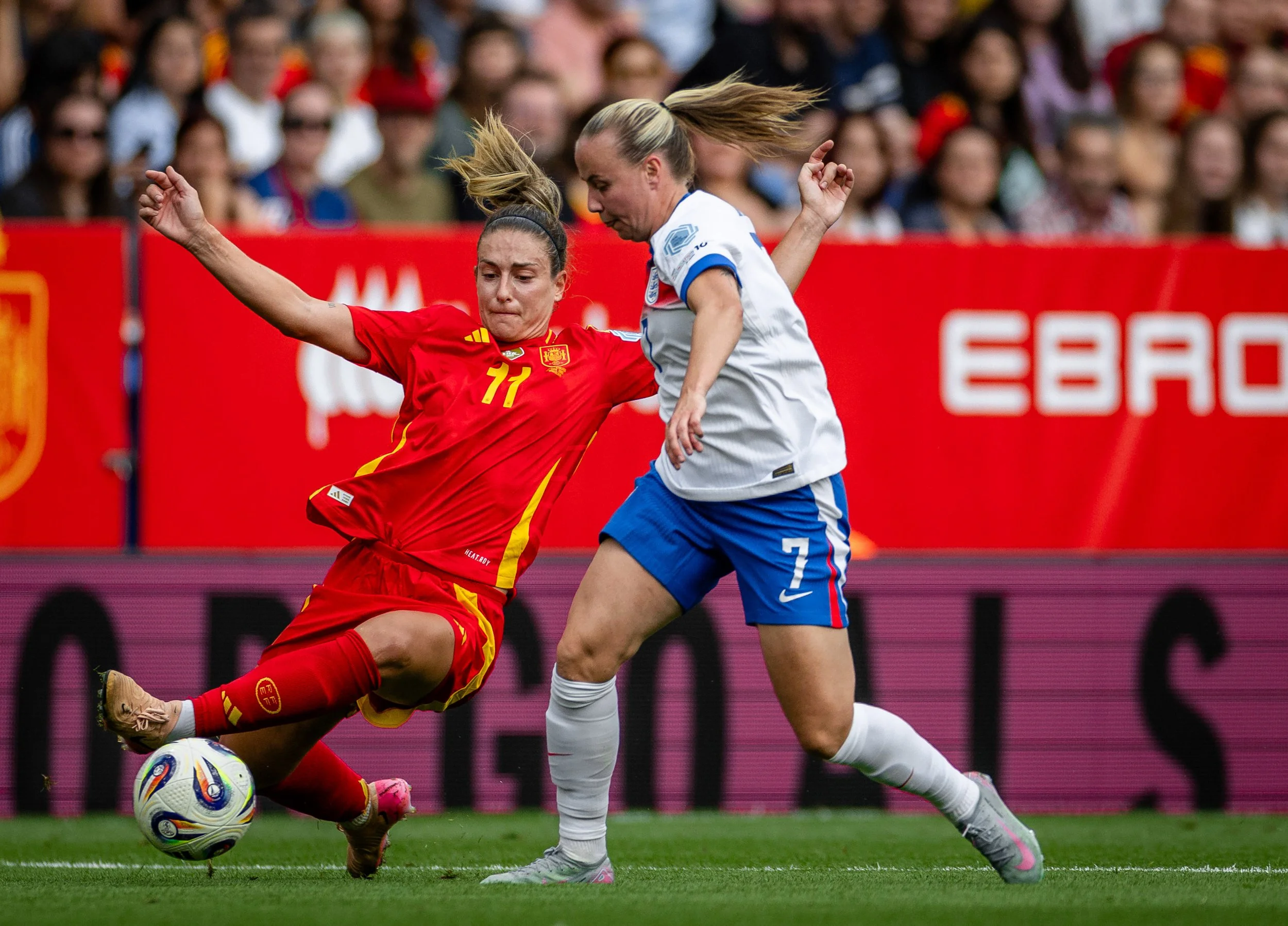 Two female soccer players, one in a red uniform and the other in a white uniform, compete for the ball during a match with a crowd of spectators in the background.