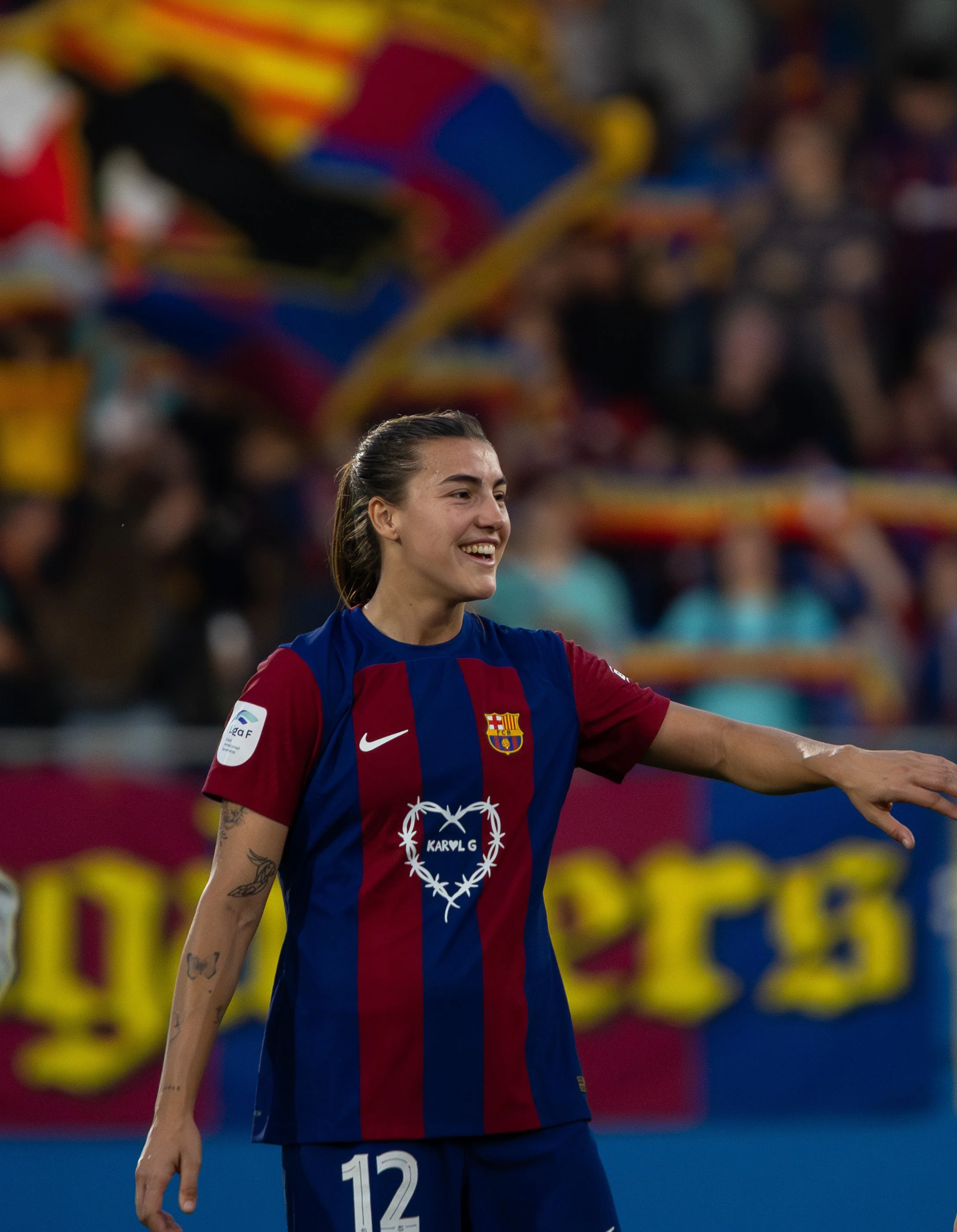 A female soccer player wearing a blue and red striped jersey with the FC Barcelona crest, the number 12 on her shorts, and a tattoo on her right arm, smiling during a match with crowd and team banners in the background.