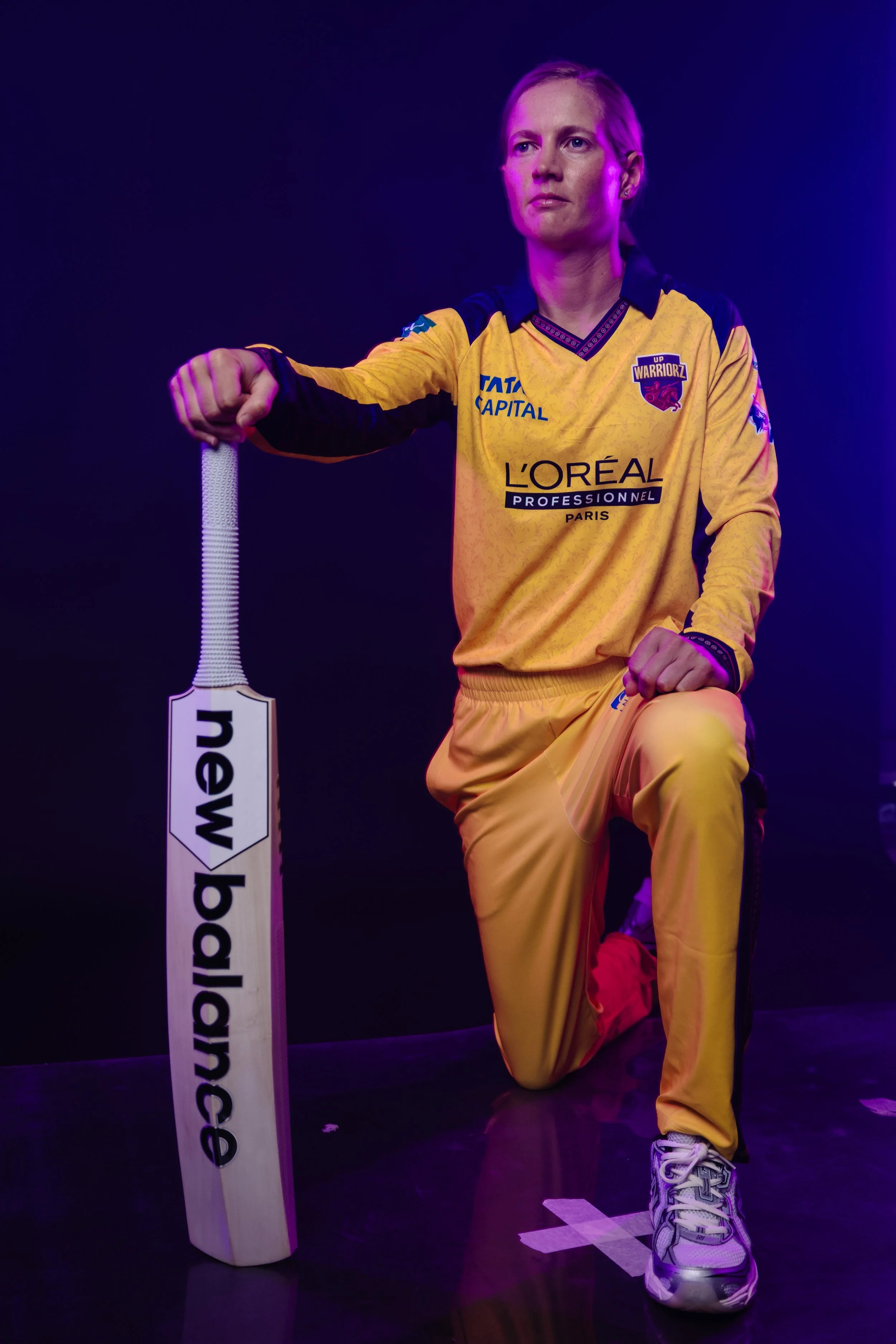 A woman in a yellow sports jersey with sponsor logos kneels on one knee, with her right hand on her knee and her left hand holding a white bat with the logo 'New Balance' on it. She is in a dimly lit setting with violet and pink lighting, wearing ath