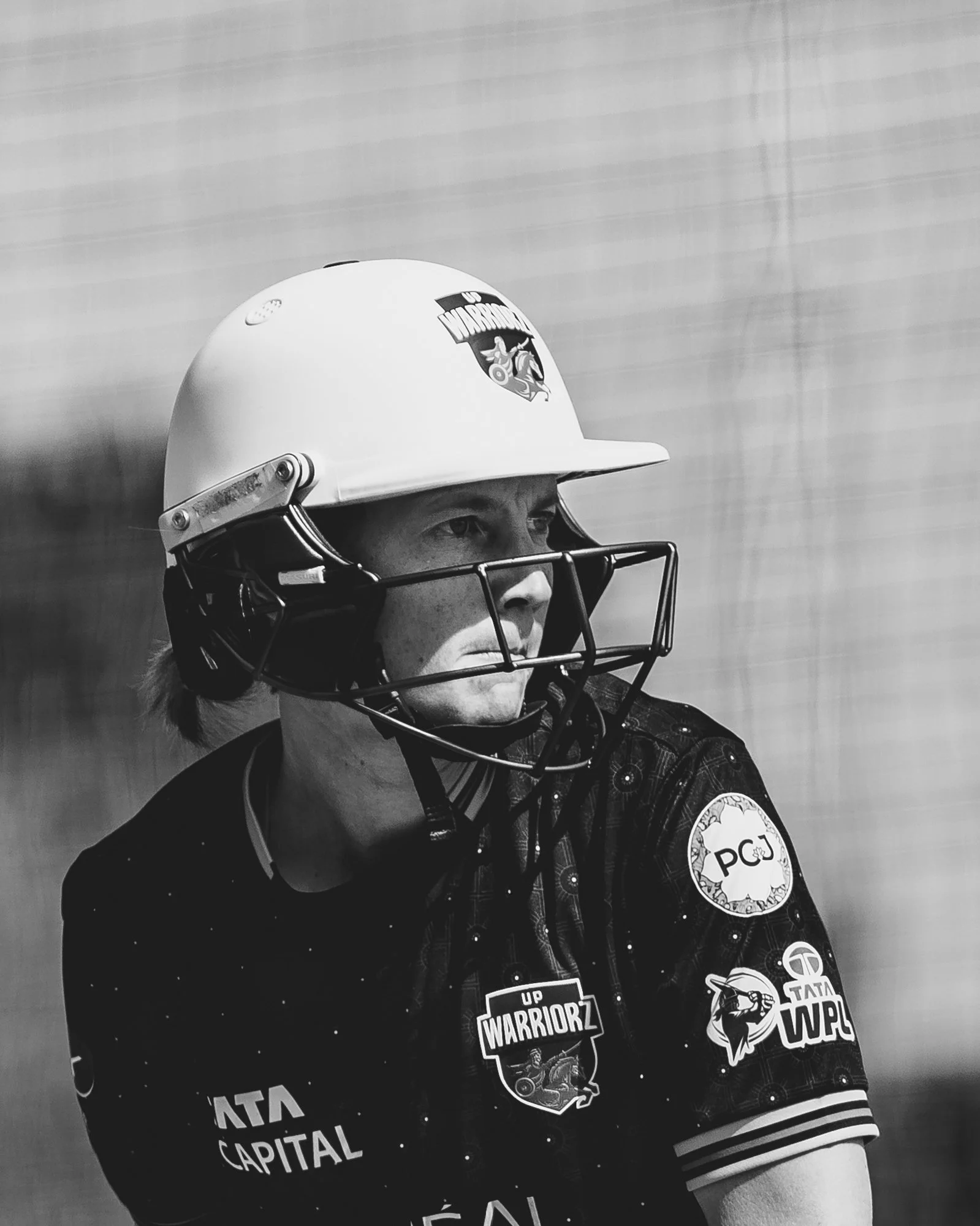 Close-up of a person wearing a helmet with the logo of the San Francisco 49ers, and a jacket with multiple patches and logos, including "UP SUPPORT WARRIORZ" and "TATA IPL".