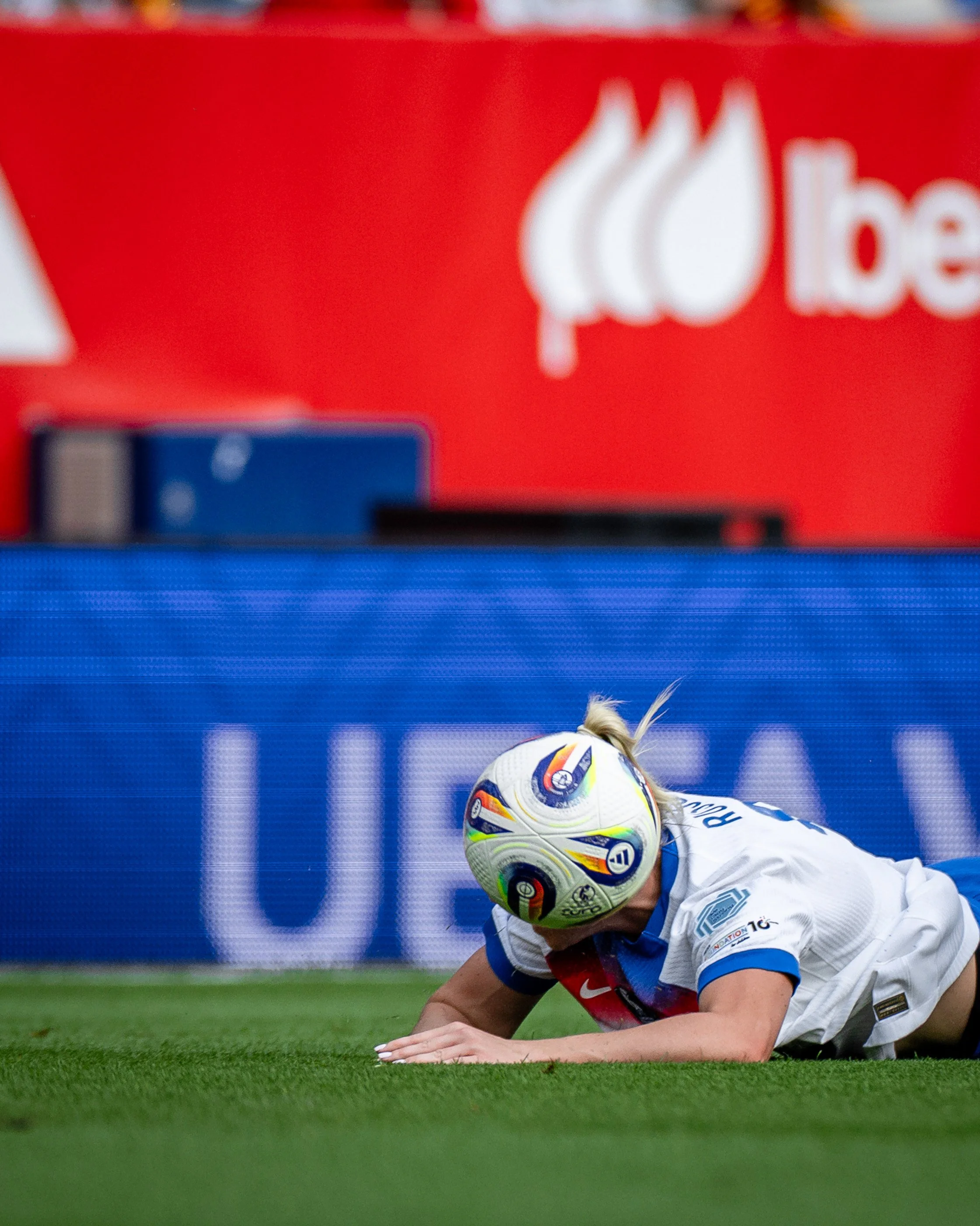 A soccer player in a white jersey is on the ground, holding a soccer ball with a rainbow-colored pattern, during a game on a green field with a digital advertising board in the background.