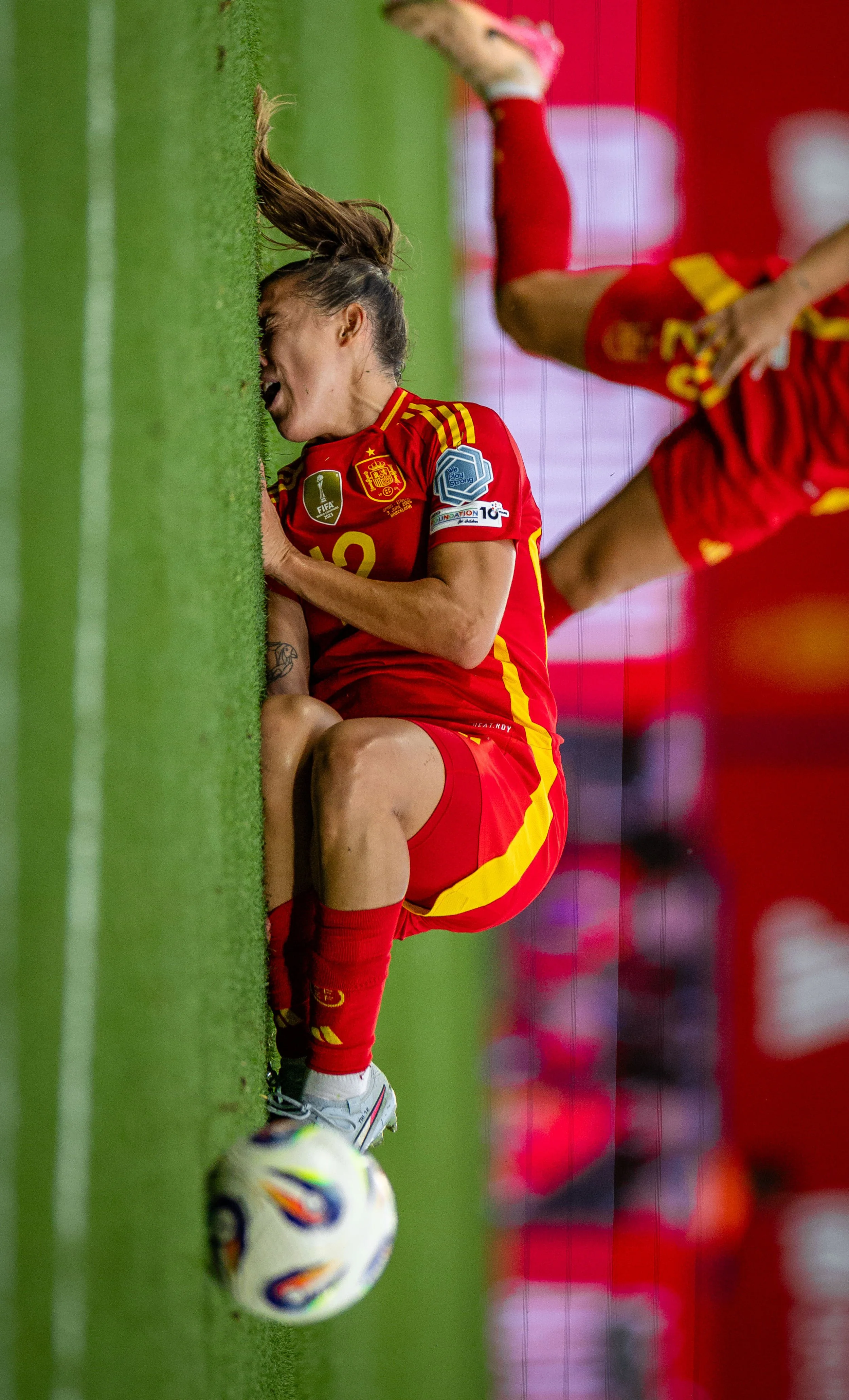 A female soccer player in a red and yellow uniform crouches on the ground, holding her leg, in pain, next to a soccer ball near the goal line.