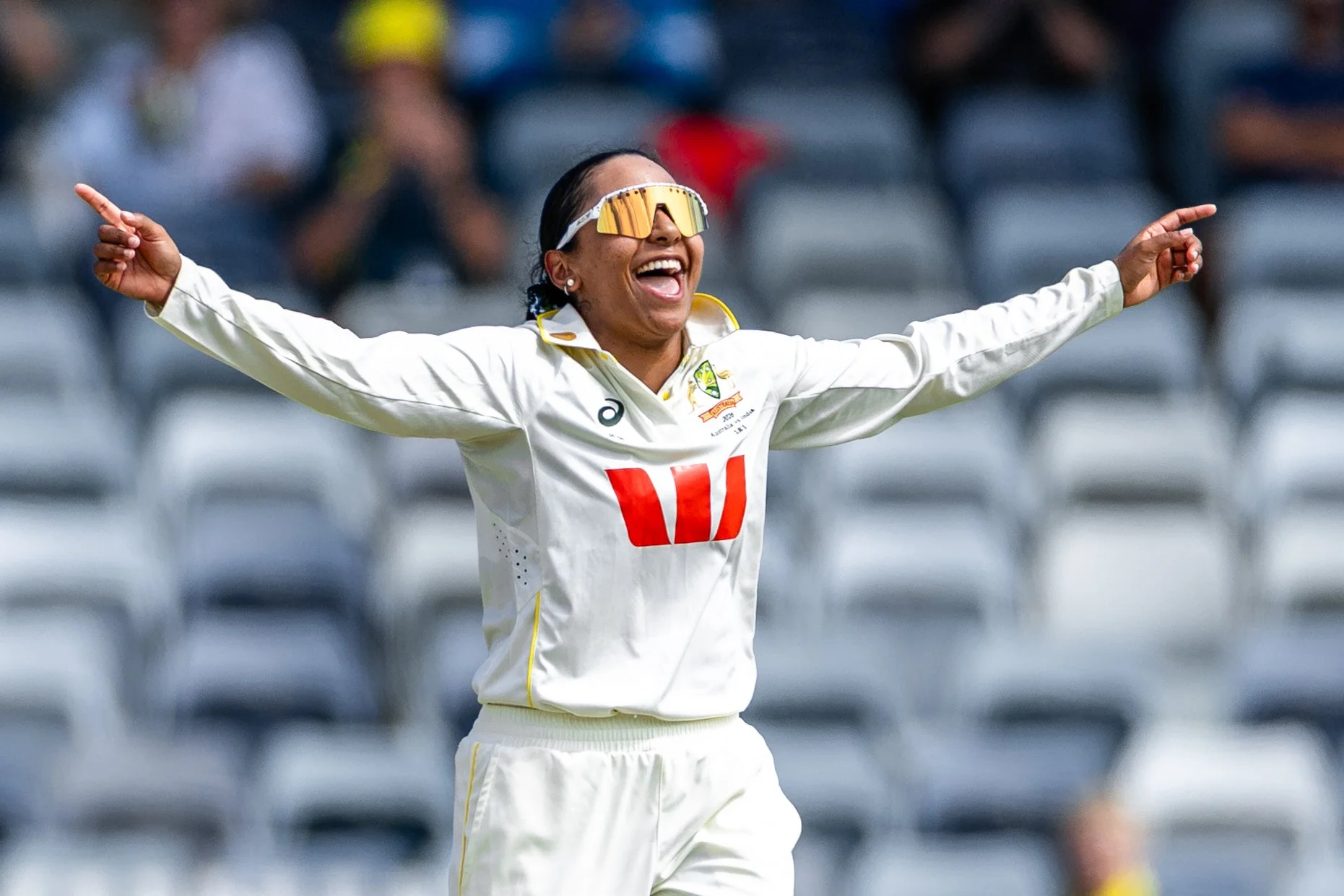 A female cricket player celebrating with her arms outstretched and a wide smile, wearing sunglasses and a white cricket uniform with yellow accents, on a cricket field.