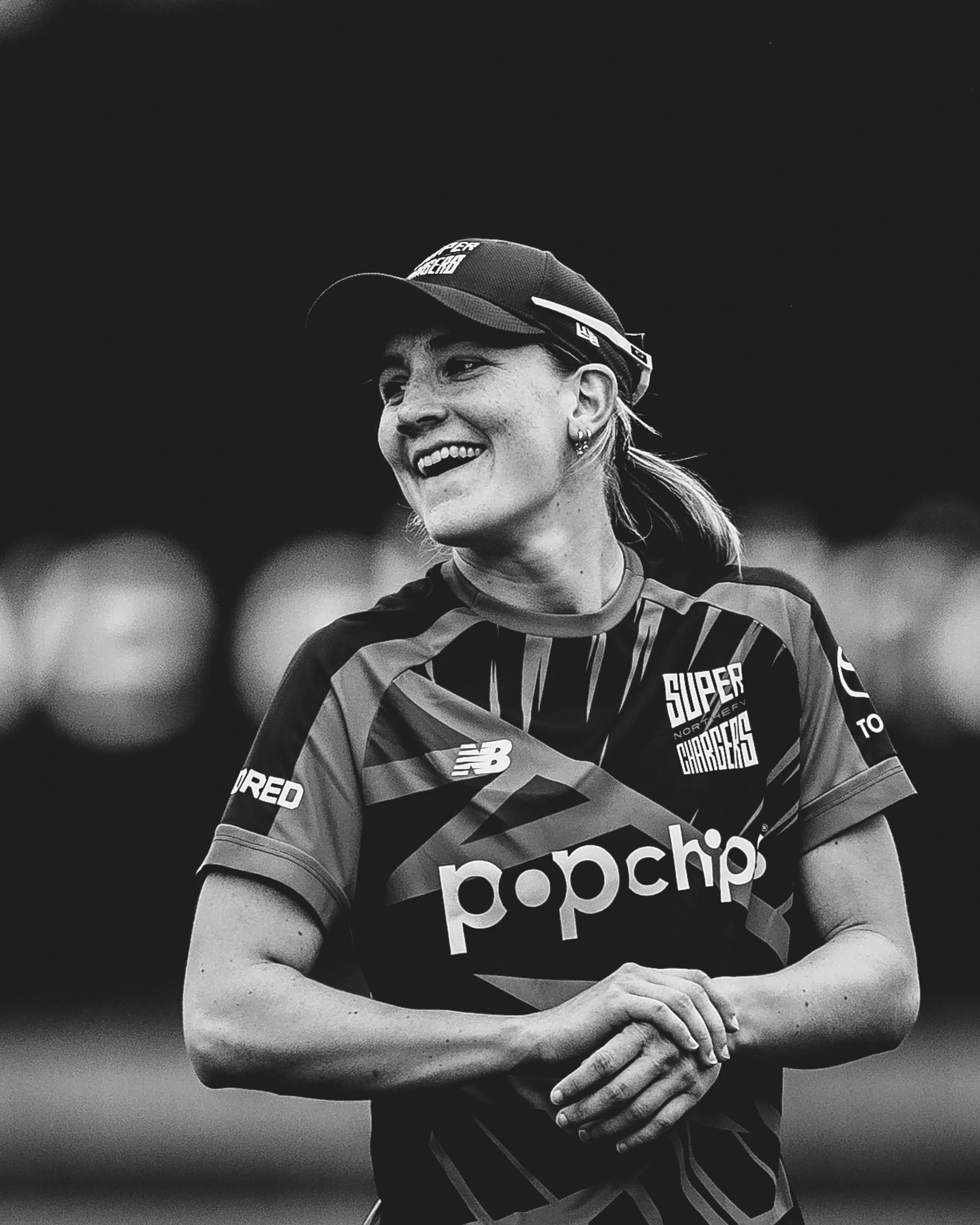 A woman smiling, wearing a sports cap and a sports jersey with the words "Super Haro" and "pocket" visible, standing against a dark background.