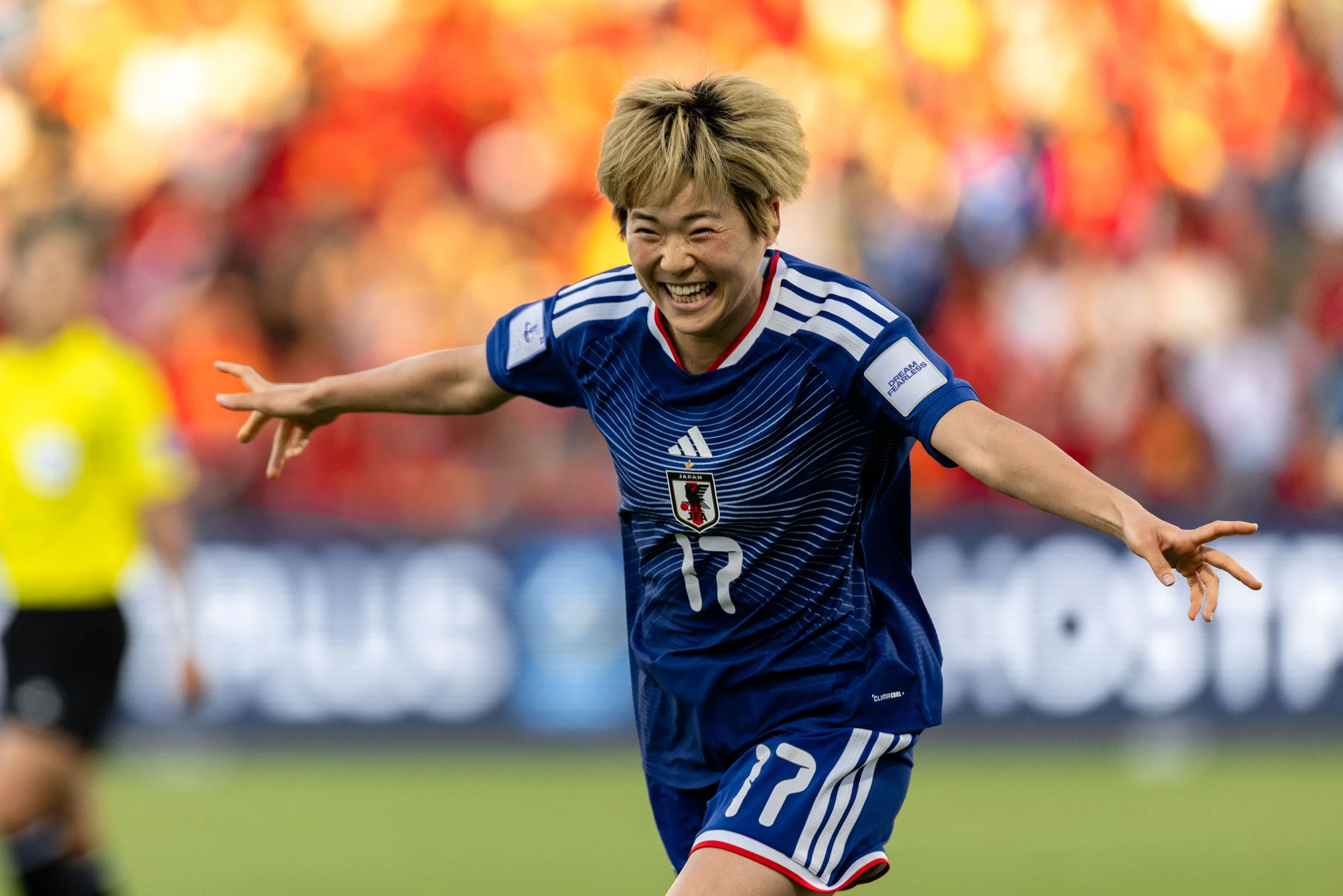 A female soccer player celebrating on the field, wearing a blue Japan national team jersey with the number 12, with a big smile and arms outstretched.