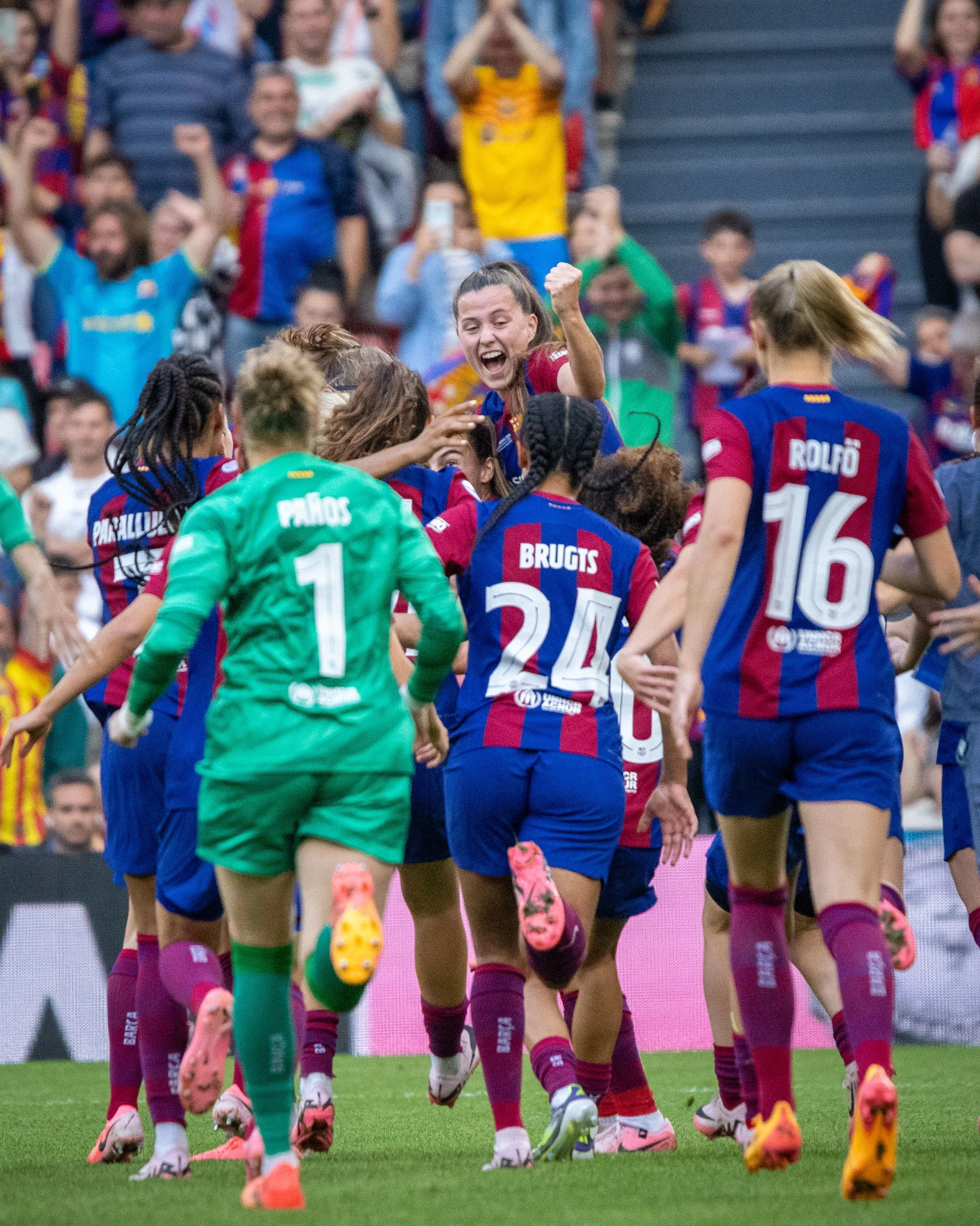 Women's soccer team celebrating on the field, with team members wearing blue and red uniforms and cheering.