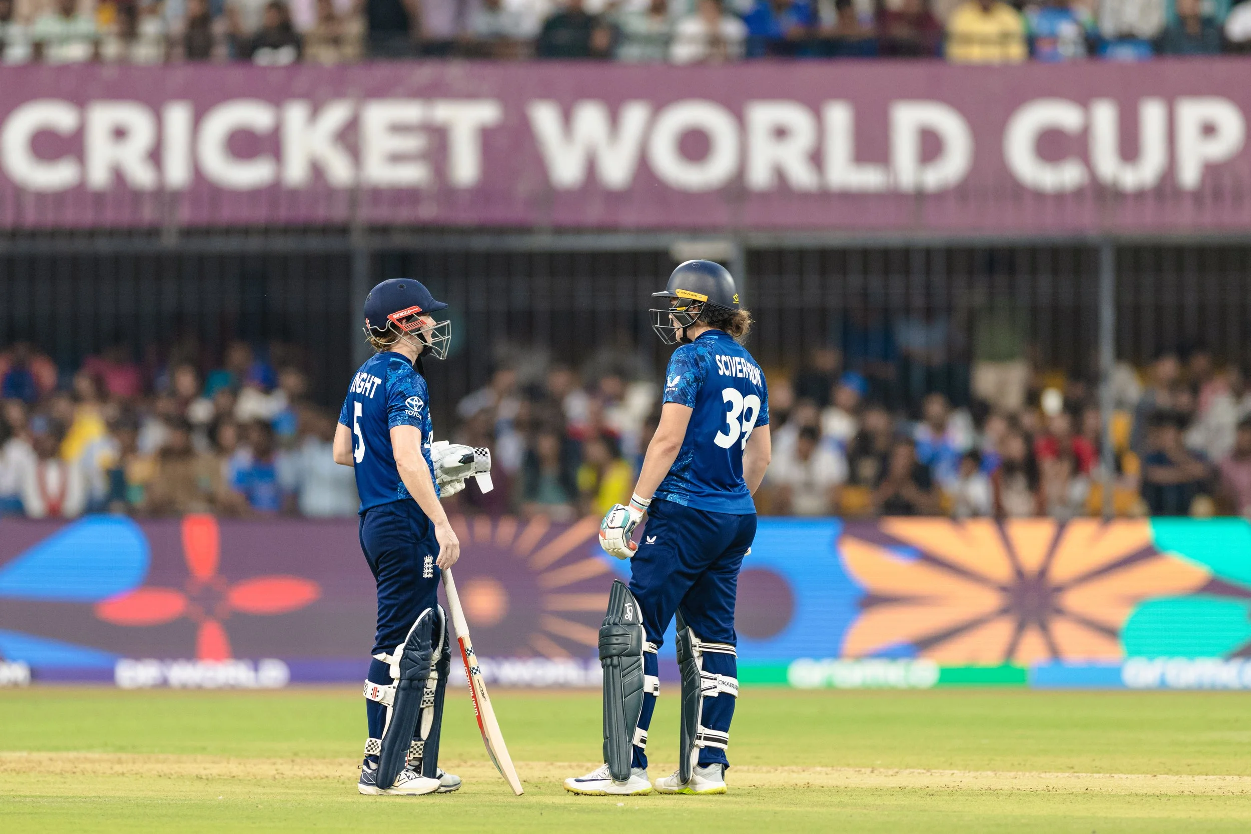 Two female cricket players in blue uniforms with helmets and pads standing on a cricket field, engaged in conversation during a match at the Cricket World Cup.