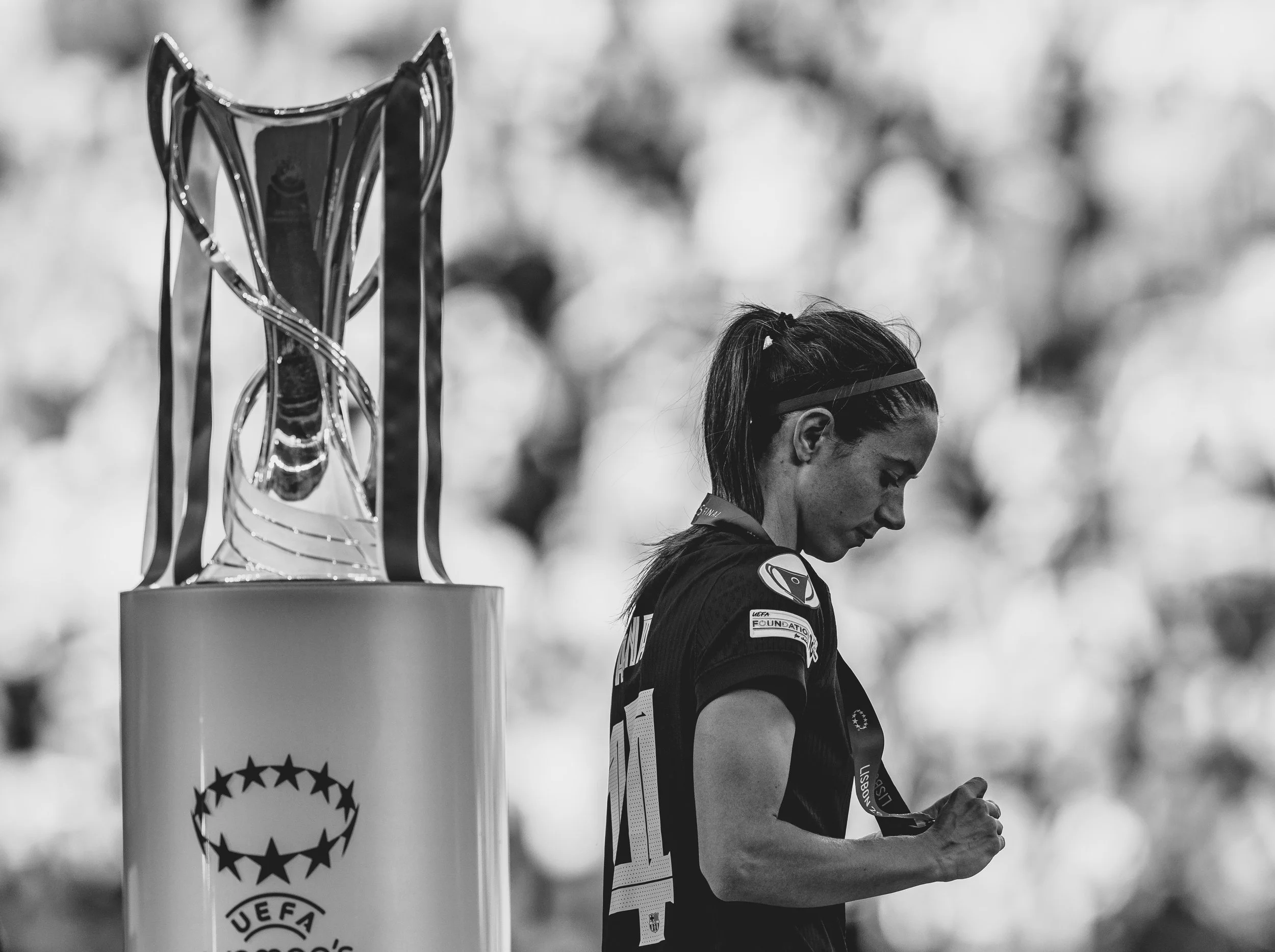 A female athlete in a sports jersey, wearing a medal, stands next to a trophy on a pedestal, looking at her phone after a competition.