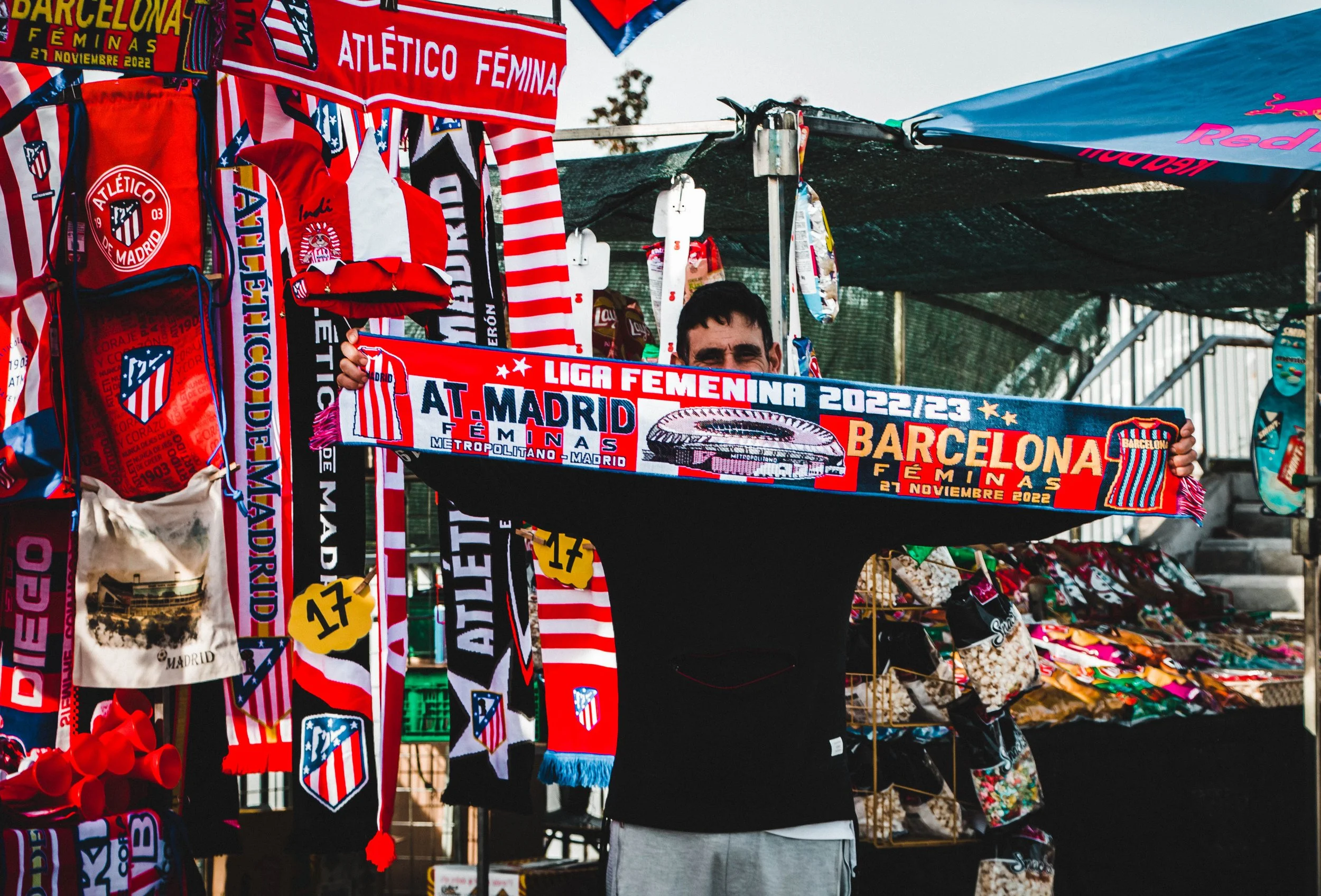 A man is holding a red, blue, and white scarf that reads "Liga Femenina 2022/23 Atletico Madrid" at a merchandise stall. The stall has various Atlético Madrid scarves, flags, and fan gear hanging on display, mainly in red, white, and blue colors, in 