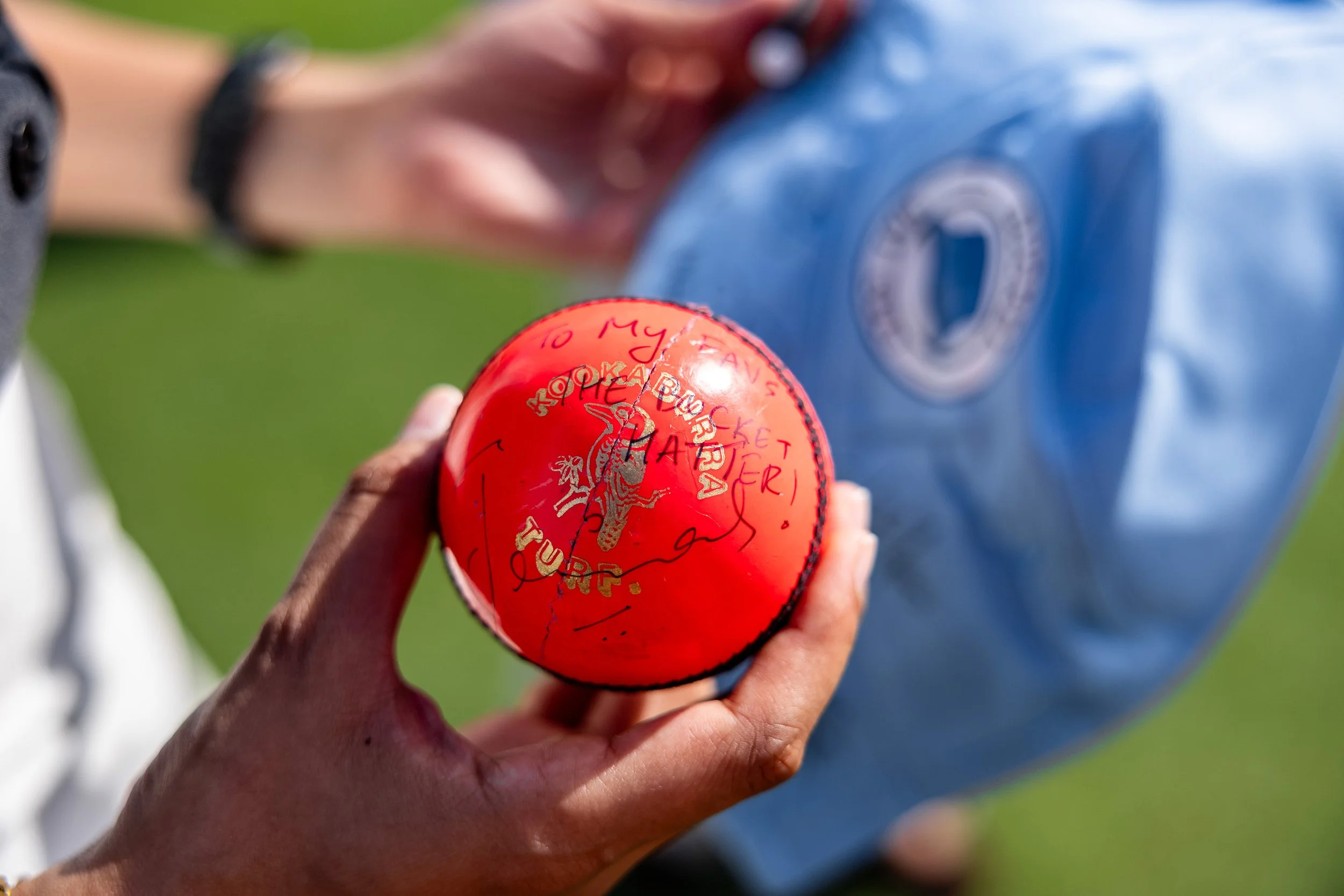 A person holding a bright red tennis ball with handwritten signatures and messages on it, with a blurry background of another person and green grass.