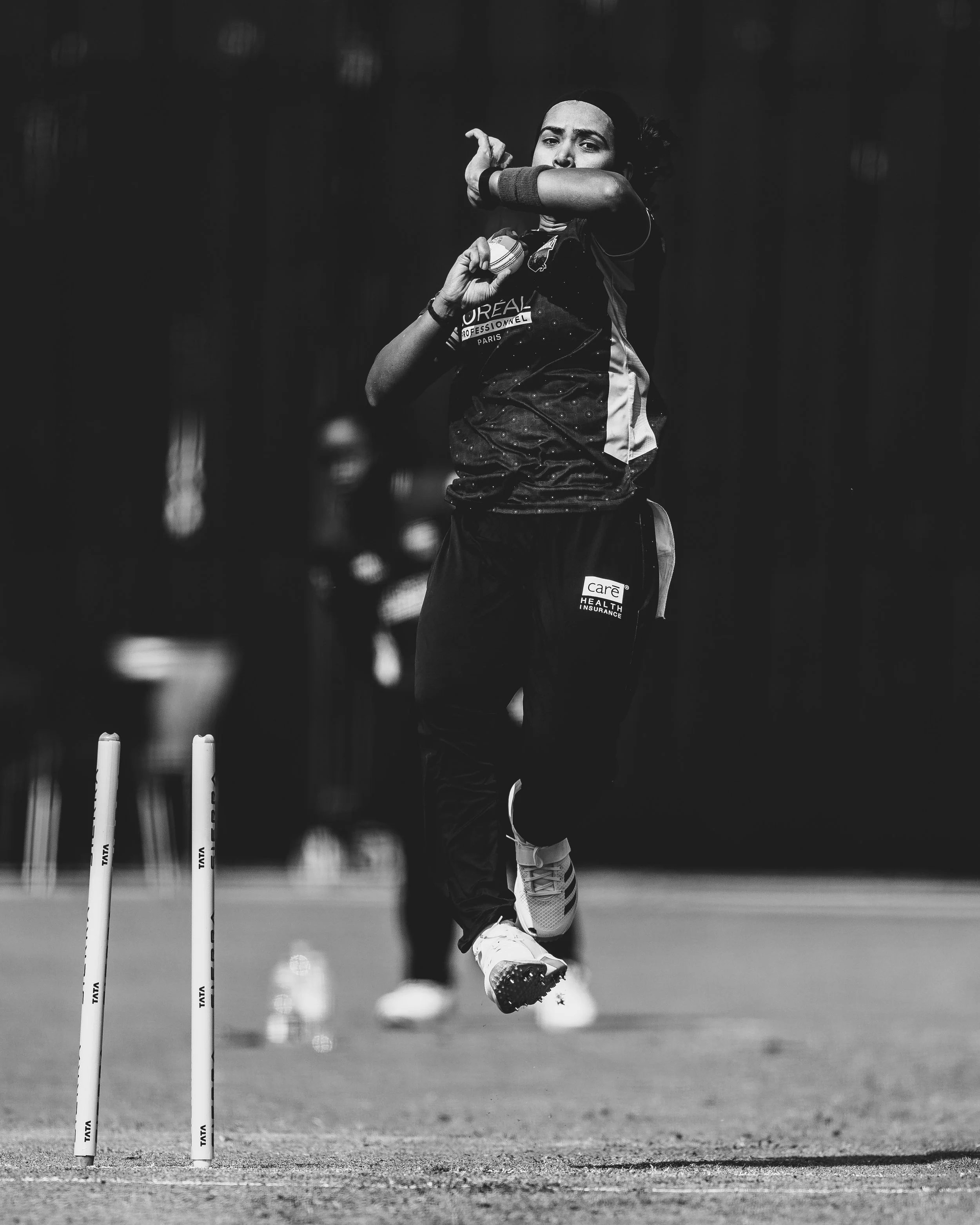 A female athlete in sportswear jumping over cricket stumps during a game on a cricket field.