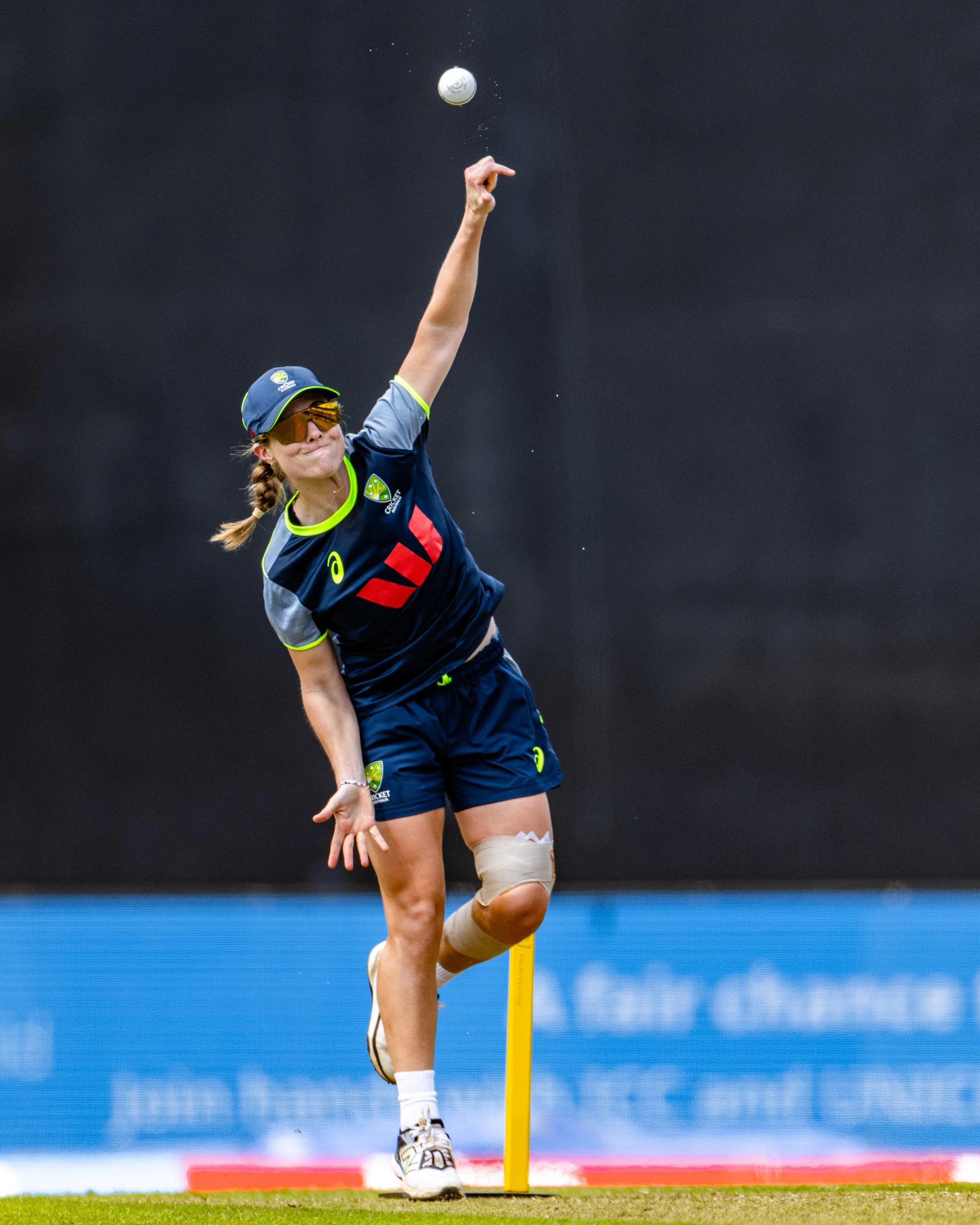 A female cricket player wearing sunglasses and a uniform, stepping over the popping crease during a cricket match, with her sweatband visible on her knee.