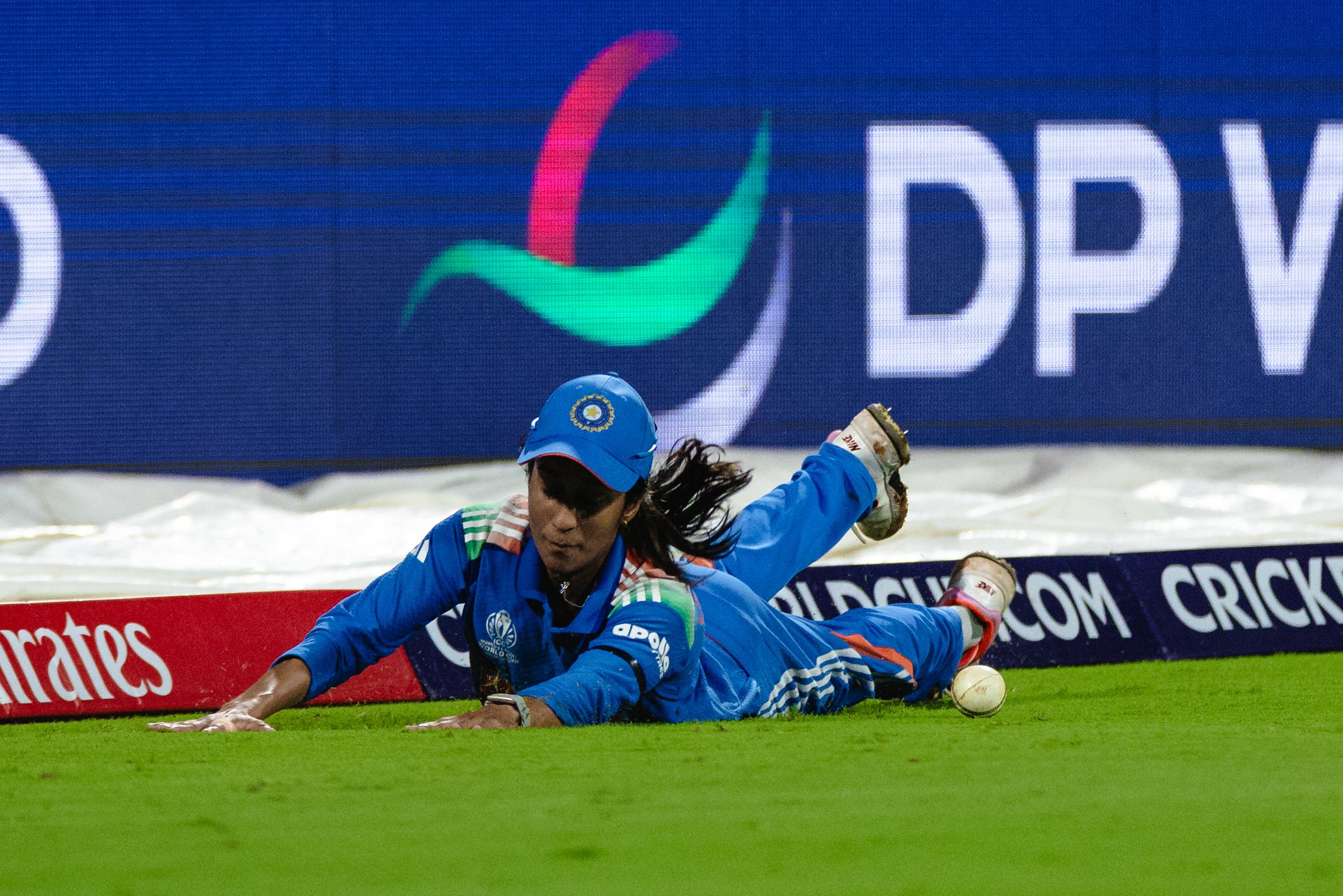 An athlete in blue sports gear, including a helmet with the Indian cricket logo, falls onto the cricket field during a match, with a cricket ball rolling on the ground nearby.