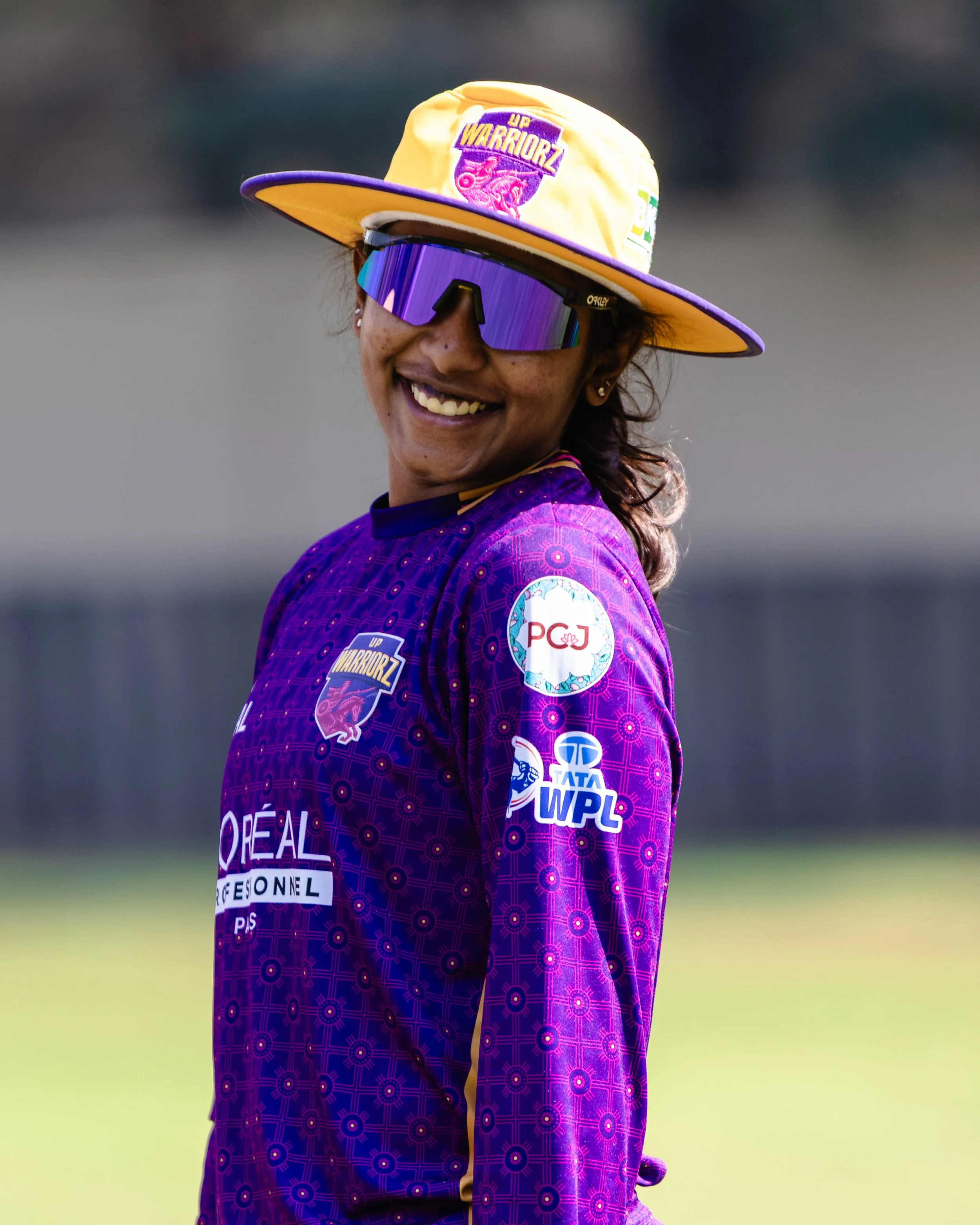 Smiling woman wearing sports jersey, large purple sunglasses, yellow hat with purple trim, and multiple sponsor patches on her sleeve and chest at a sporting event.