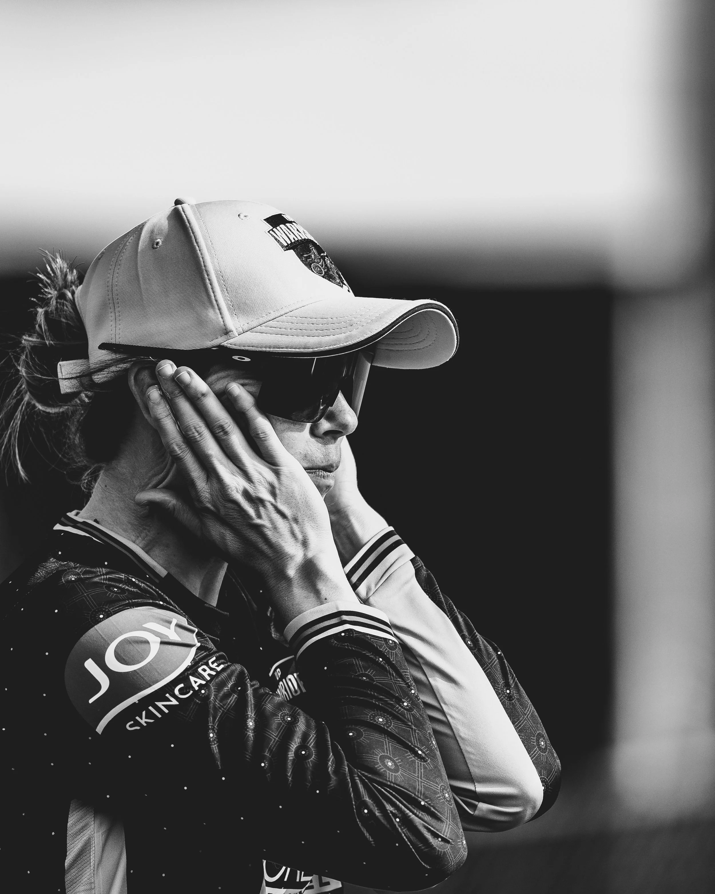 A woman wearing a baseball cap, sunglasses, and a sports jacket, with her hand on her headphones, appears to be enjoying music or a moment of reflection.