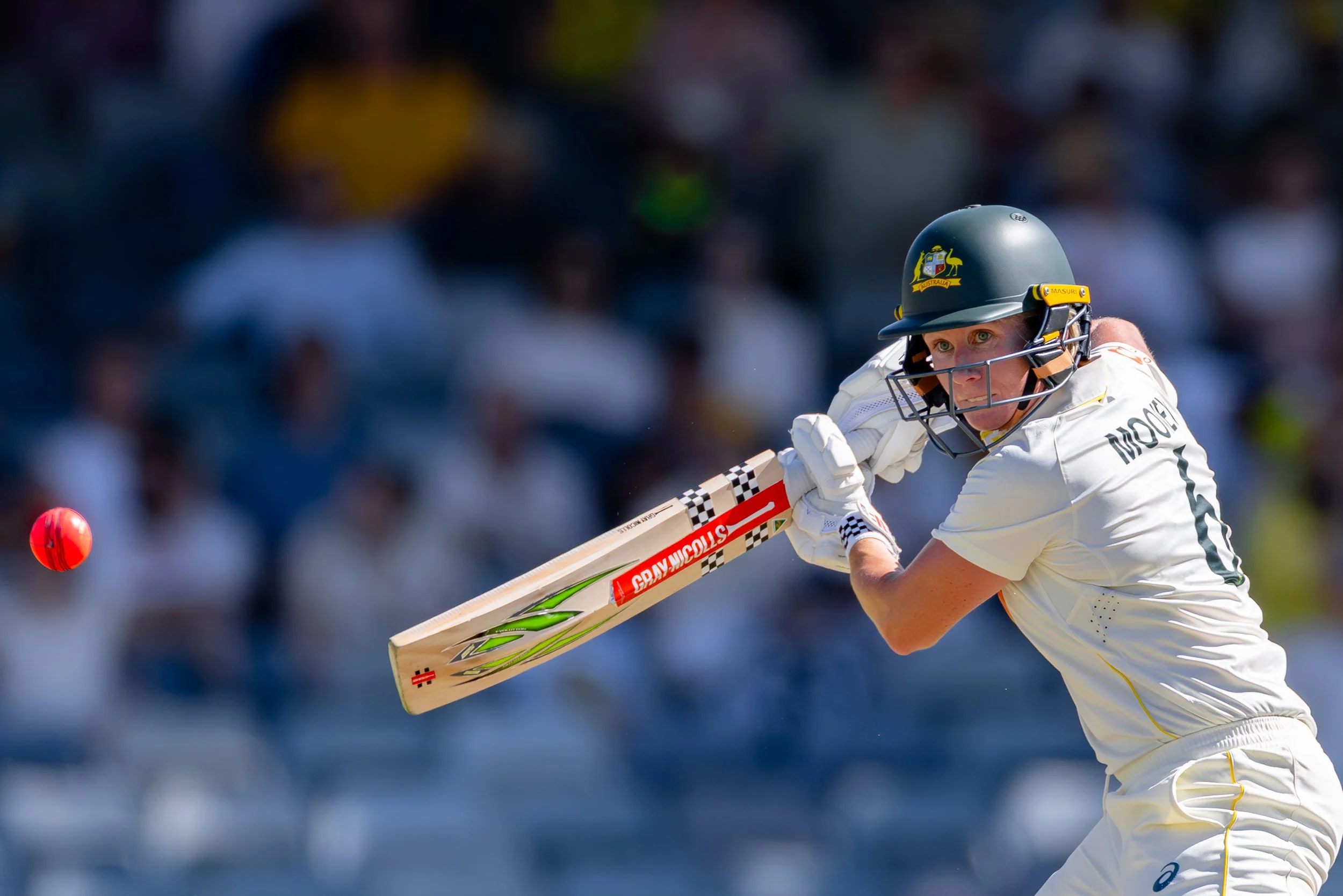 A female cricket player is in action, wearing a helmet and white uniform, hitting a ball with her bat during a cricket match.