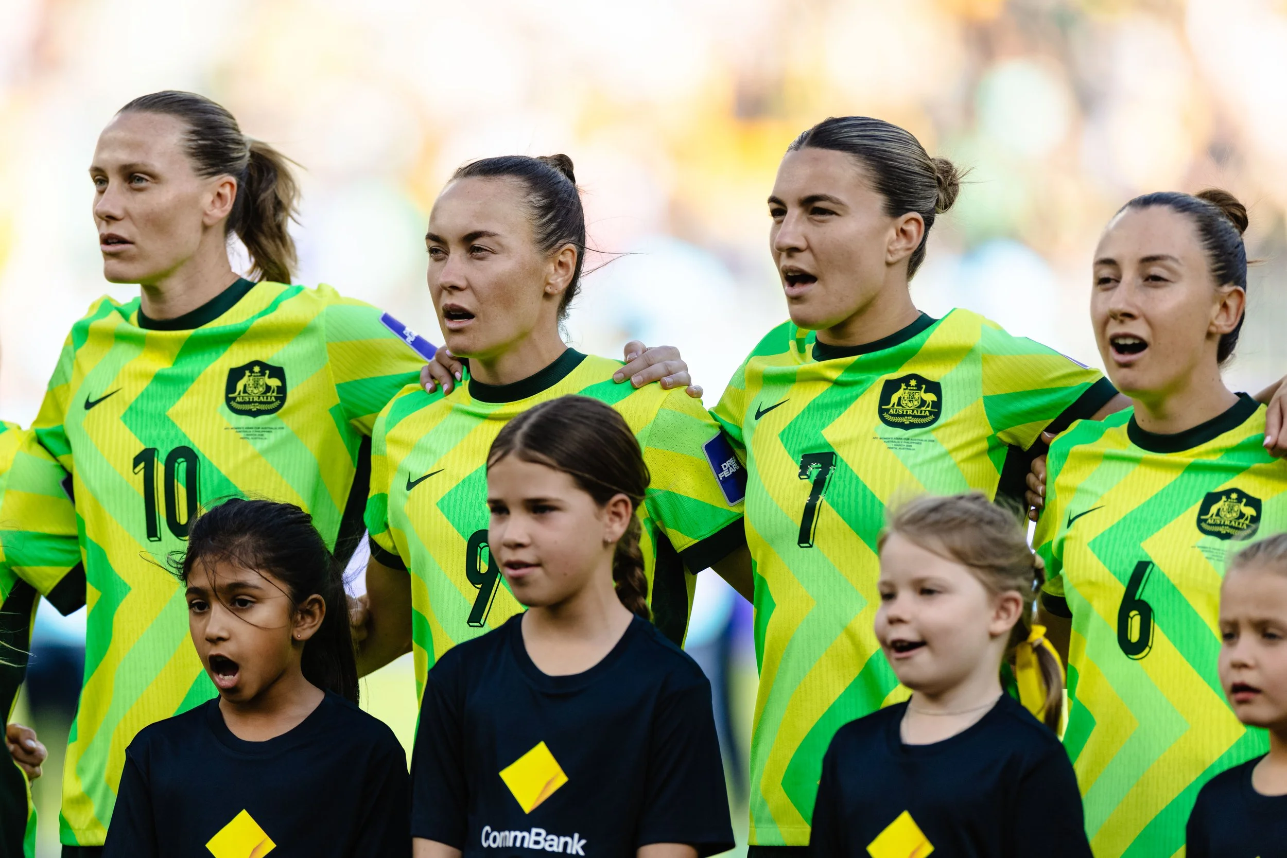 Australian women's national soccer team standing in a line with young children in black jerseys during the national anthem before a match, with players wearing yellow and green uniforms with the Australian emblem.