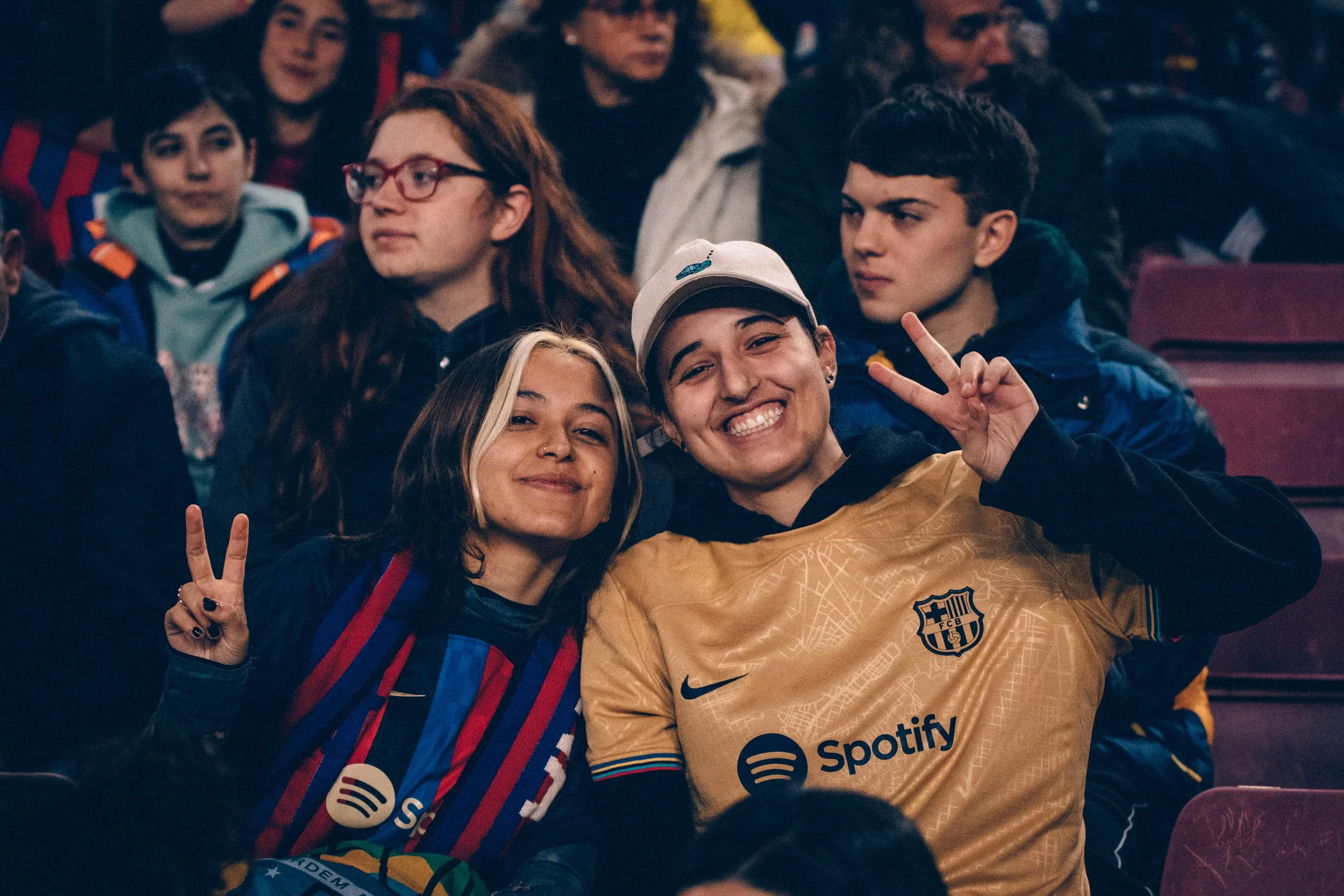 Group of young people at a sporting event, two in the front smiling and making peace signs, wearing football jerseys and casual clothing, seated in a stadium.