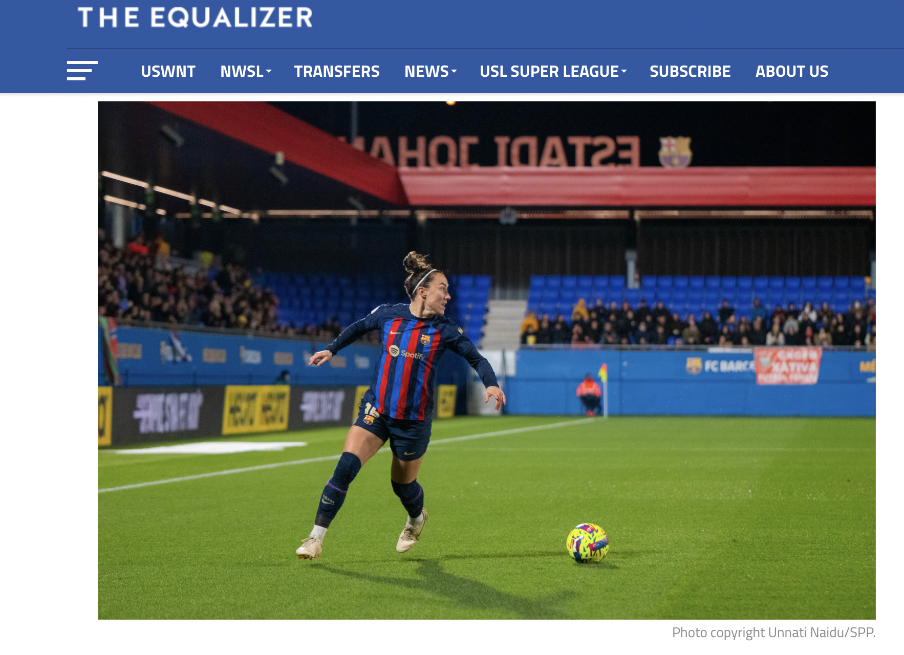 A female soccer player in a FC Barcelona uniform kicks a colorful soccer ball on the field during a match at night. There are spectators in the stands and stadium advertising boards visible in the background.