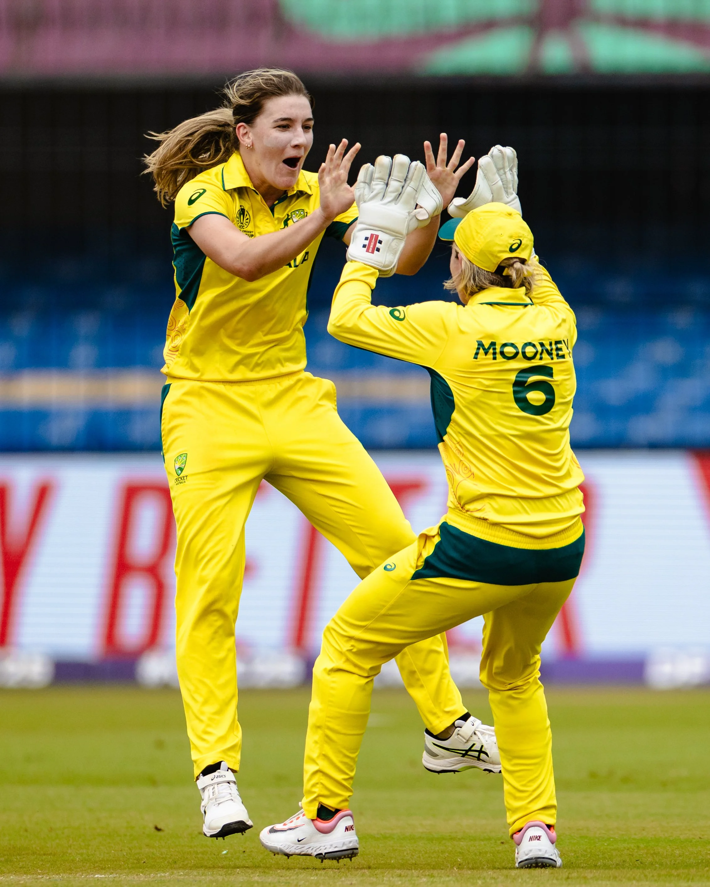 Two female cricket players in yellow uniforms celebrating on the field, with one jumping and the other with hands raised for a high-five.