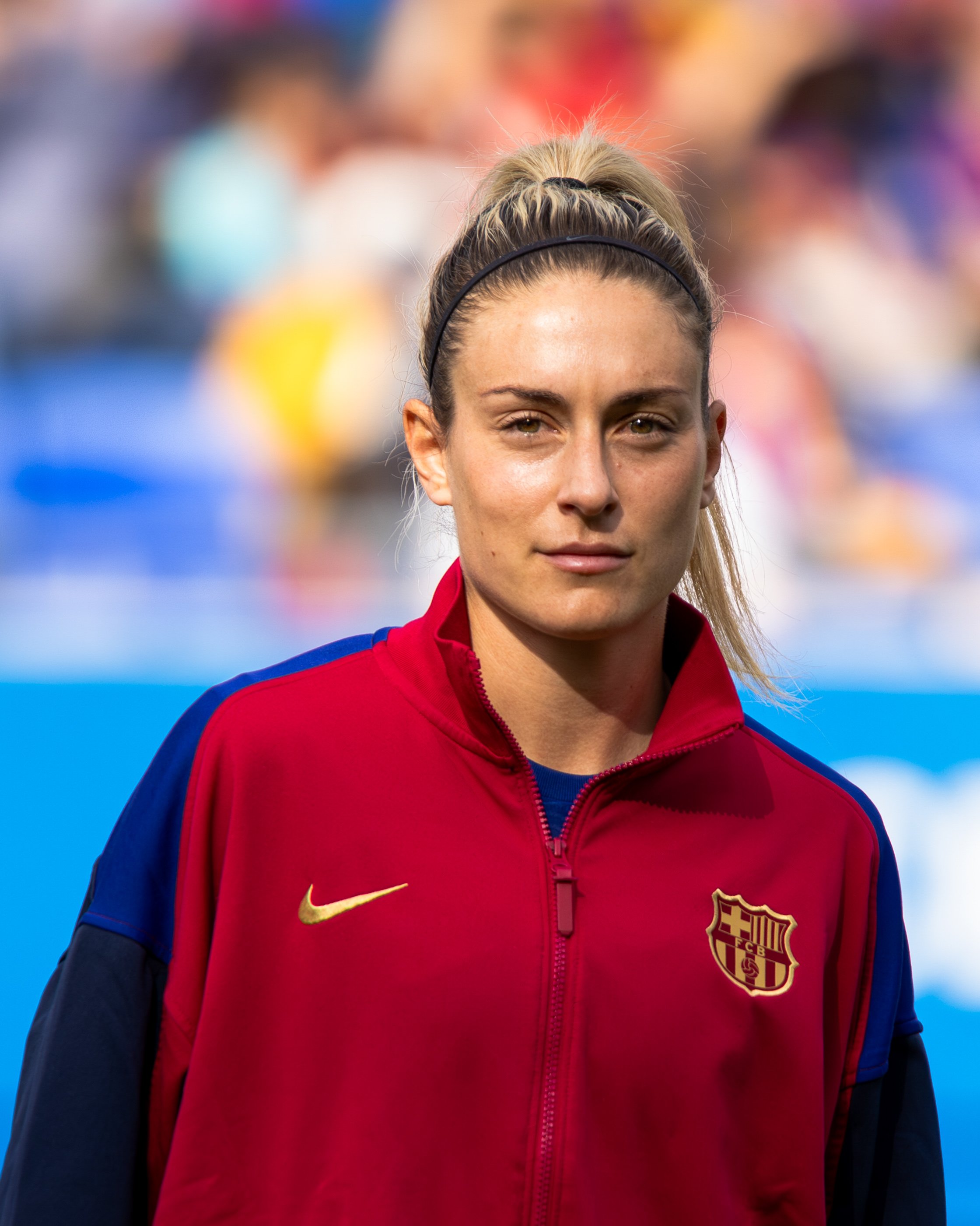 Close-up of a female athlete wearing a red and blue sports jacket with FC Barcelona logo, and a black headband, standing outdoors with a blurred crowd in the background.