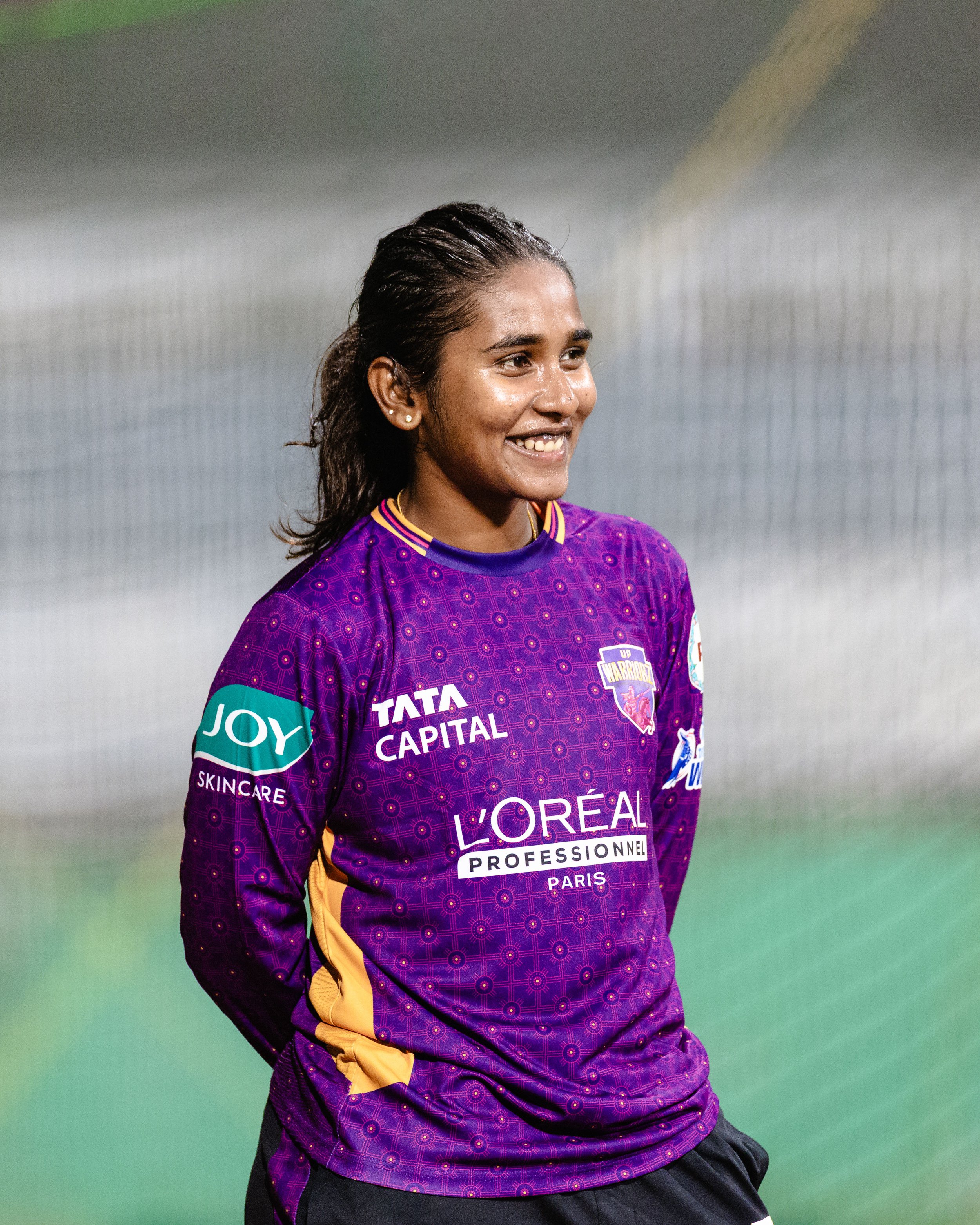 Close-up of a happy woman with dark, wet hair, wearing a purple sports jersey with sponsorship logos, standing on what appears to be a sports field.