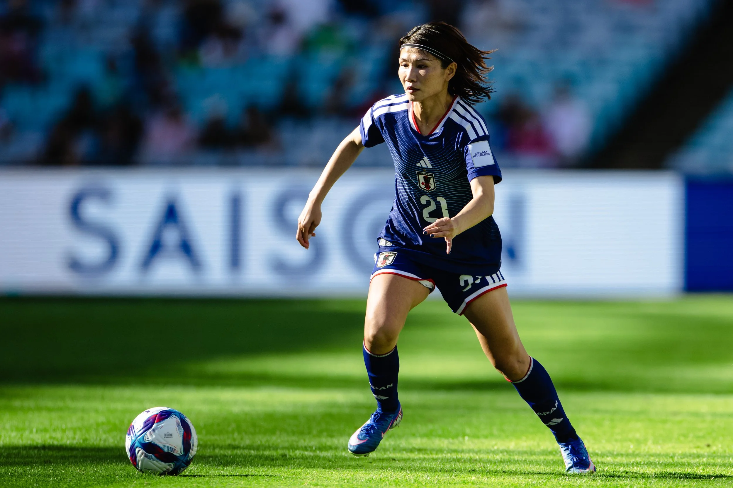 A female soccer player in a blue jersey with the number 20 on it, running on a green field and preparing to kick a soccer ball.