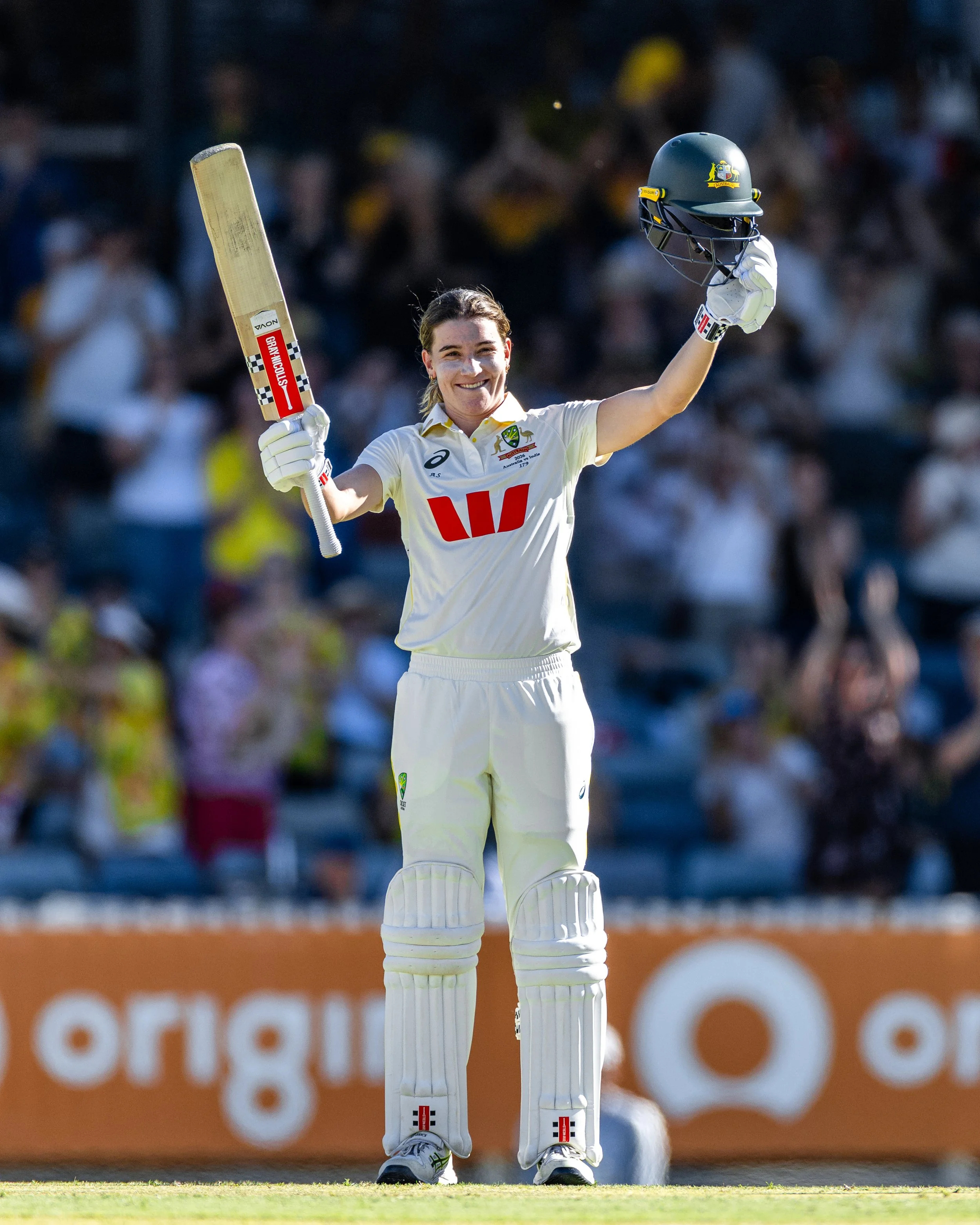 A female cricket player celebrating on the field, holding her cricket helmet in her right hand and her bat raised in her left hand. She is dressed in white cricket gear and standing in front of an audience in the stands.